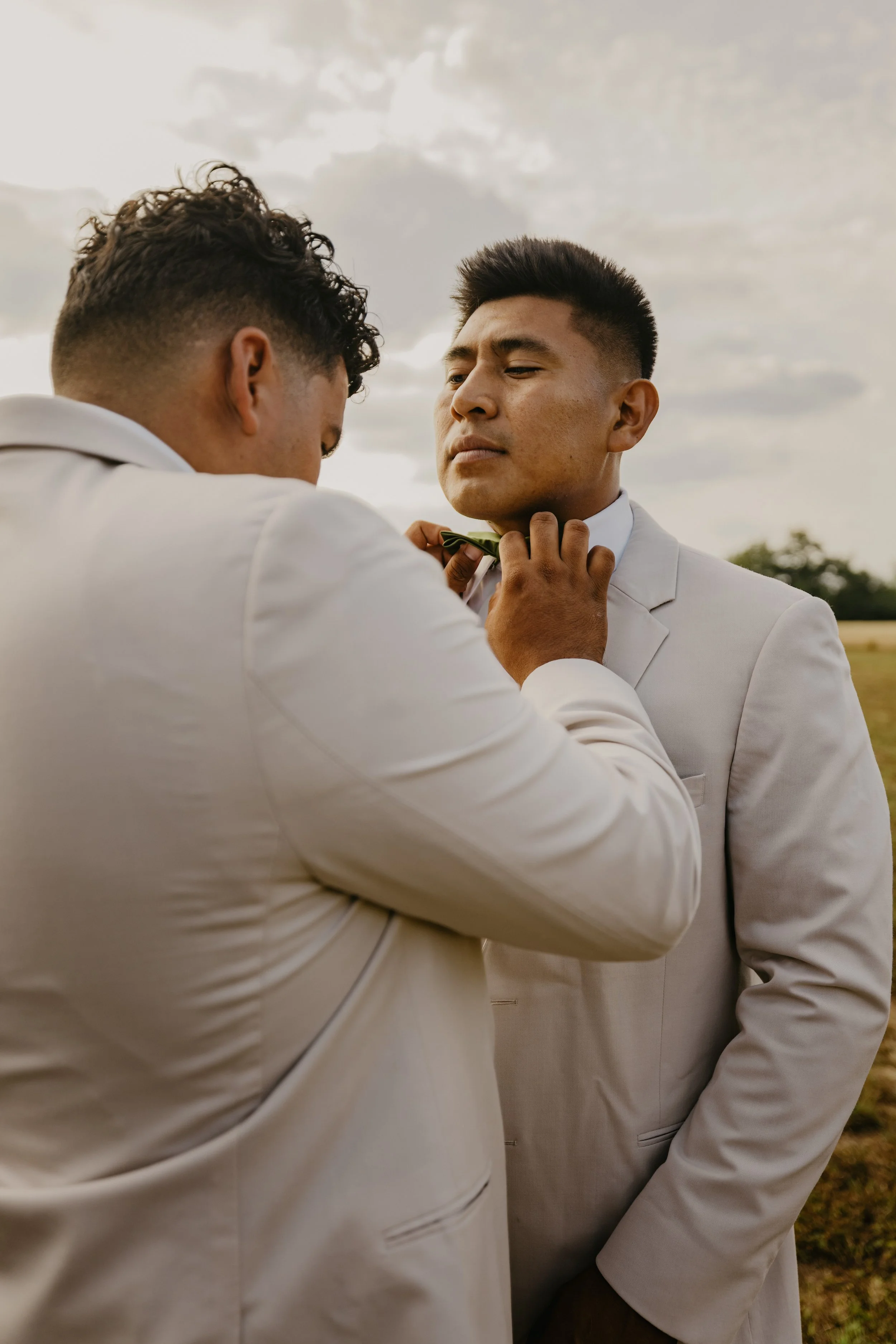 Two men in white suits outdoors, one helping the other adjust his tie, with cloudy sky in the background.