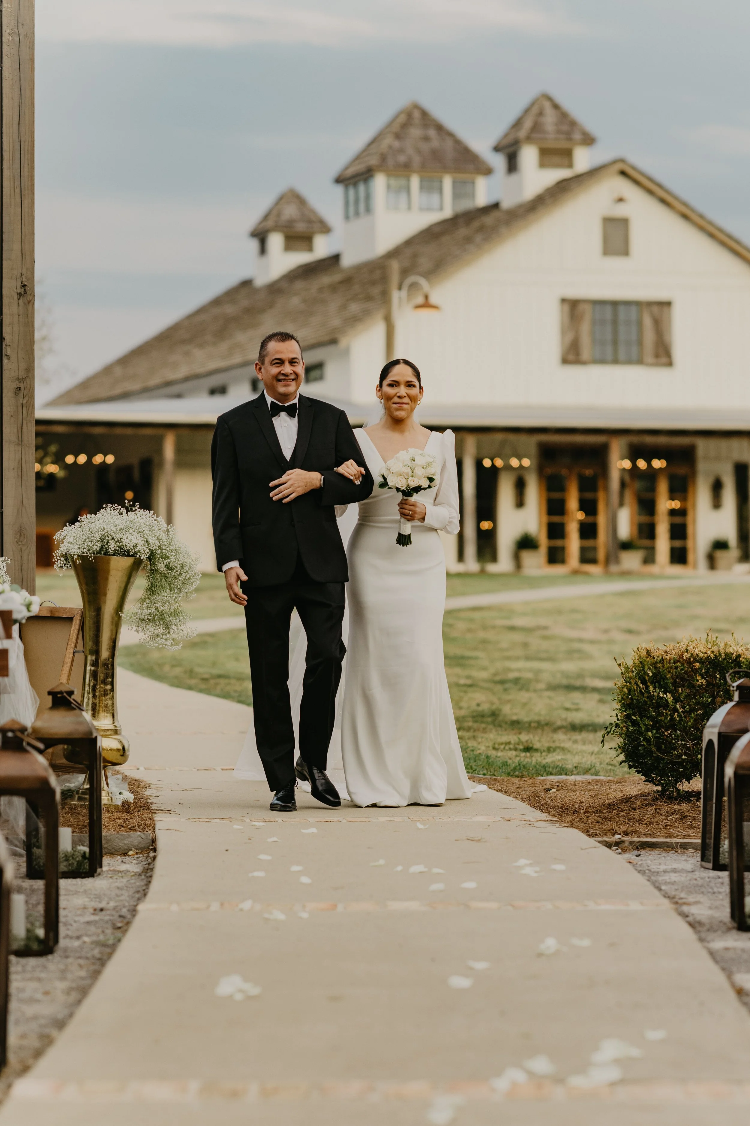 Bride walking down the aisle with her father at an outdoor wedding venue, holding a bouquet of white roses, in front of a large white barn-style building with a rustic design.