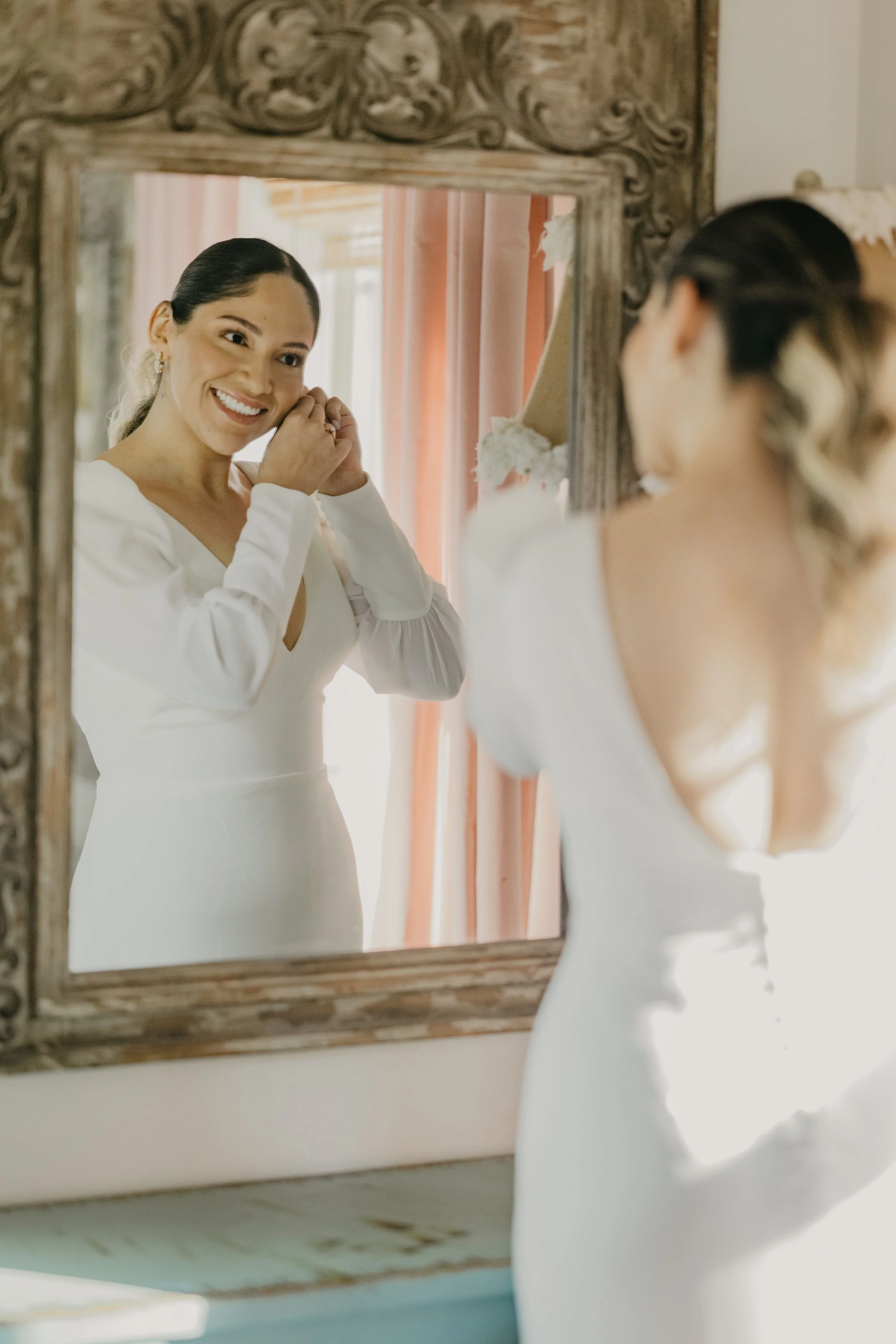 A woman in a white dress, smiling, adjusting her earring while looking at her reflection in an ornate wooden mirror.