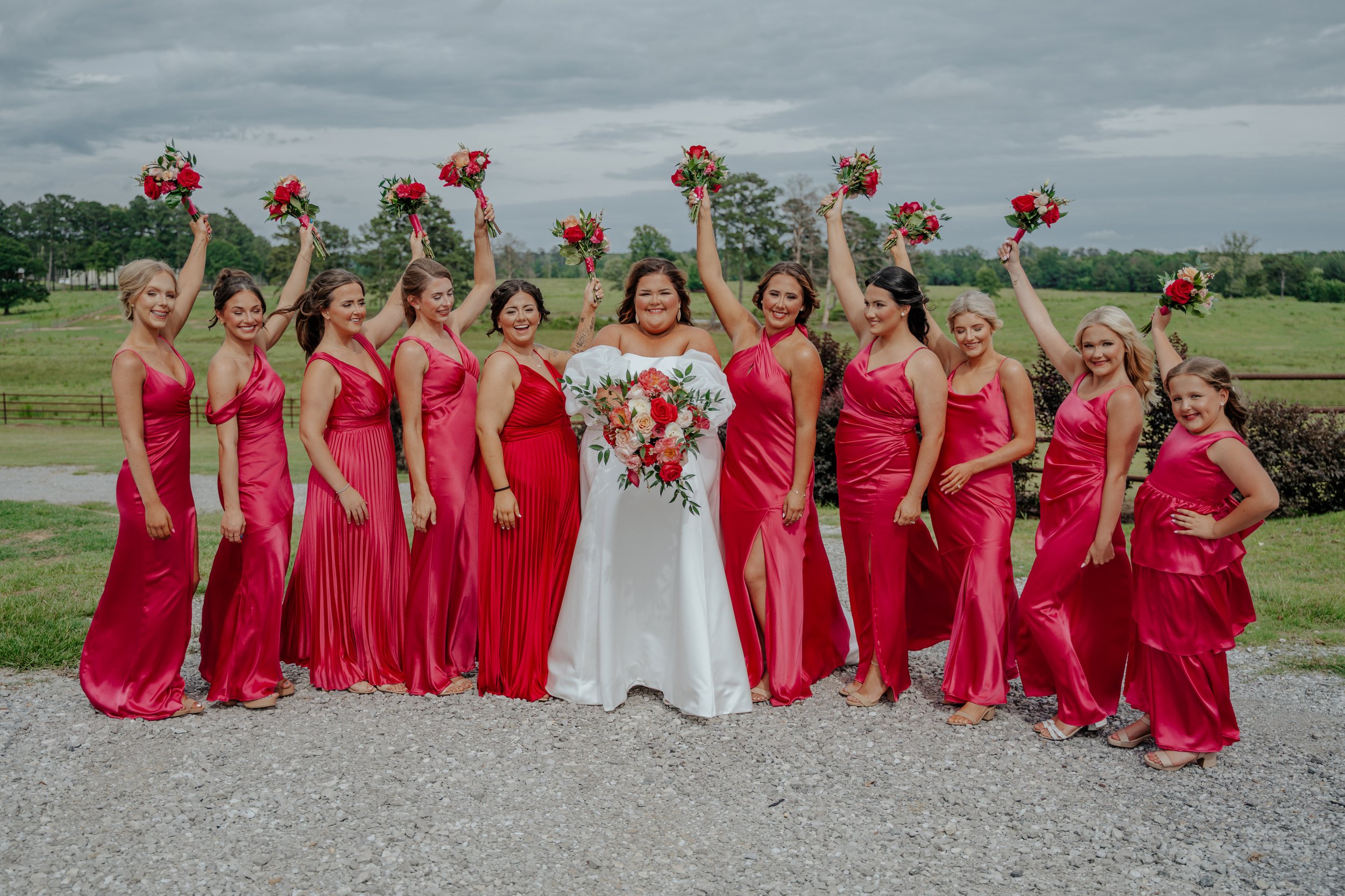 A group of women in pink dresses celebrating at a wedding outdoors, with the bride holding a bouquet and others raising smaller bouquets in the air.