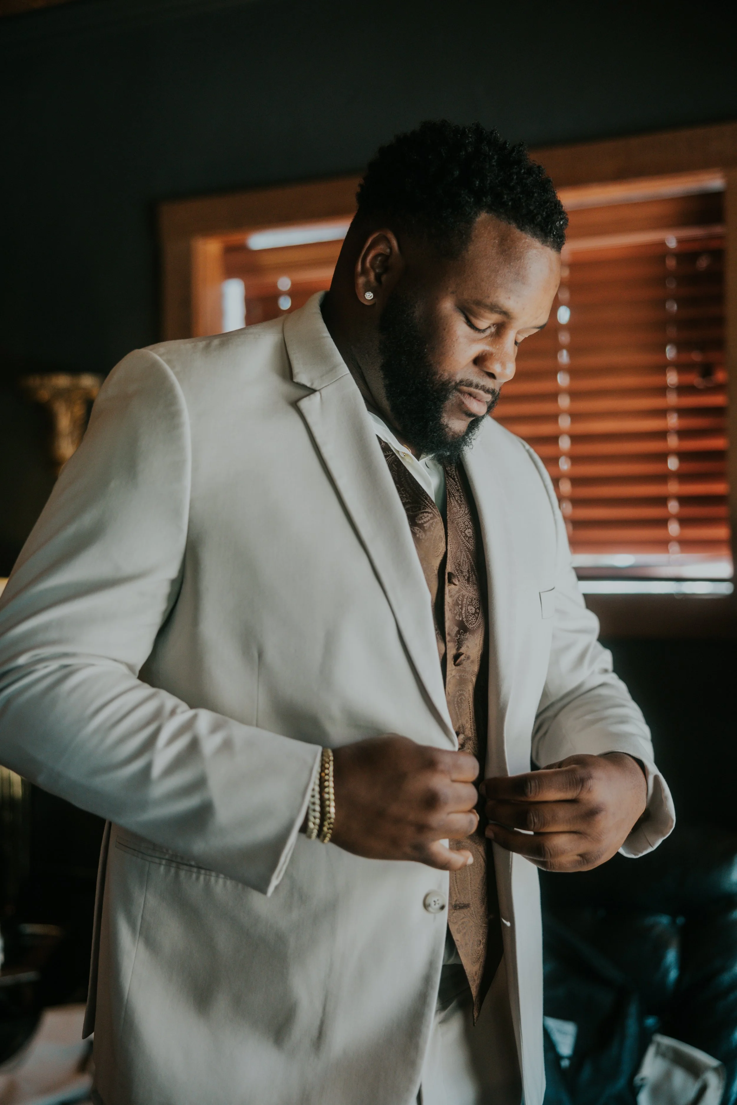 A man in a light-colored suit adjusting his jacket indoors near a window with wooden blinds.