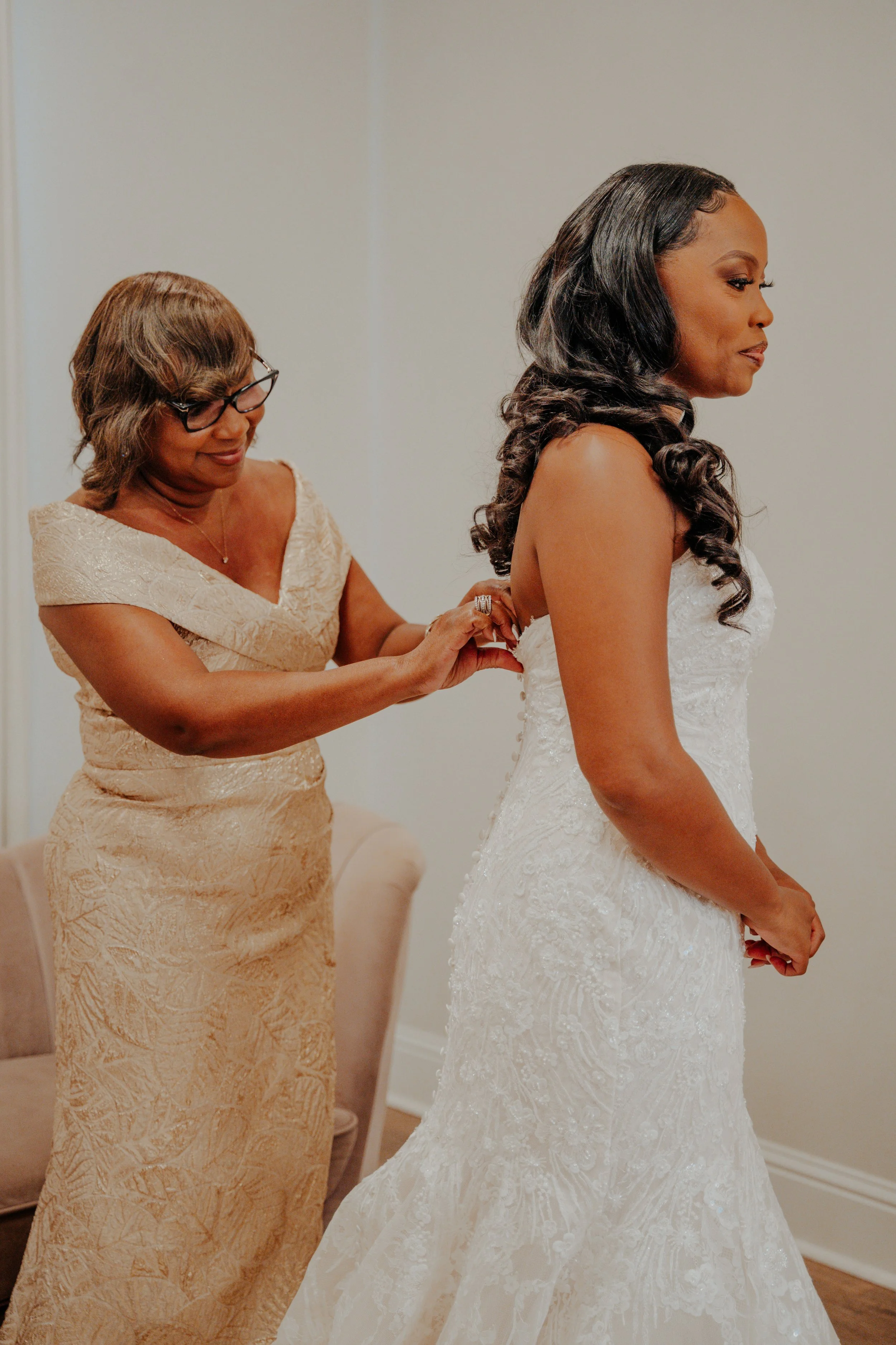 A woman in a wedding dress is being helped by another woman, likely her mother, as she prepares for her wedding.