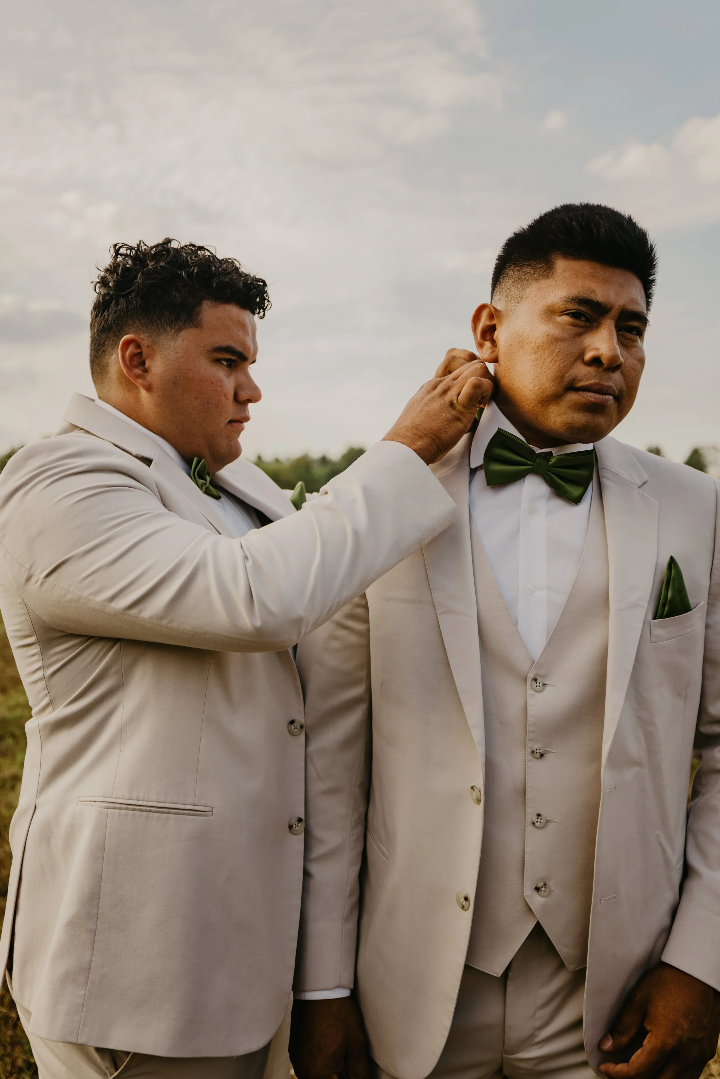 Two men in beige suits with green bow ties are outdoors, one adjusting the other's collar, with a cloudy sky in the background.