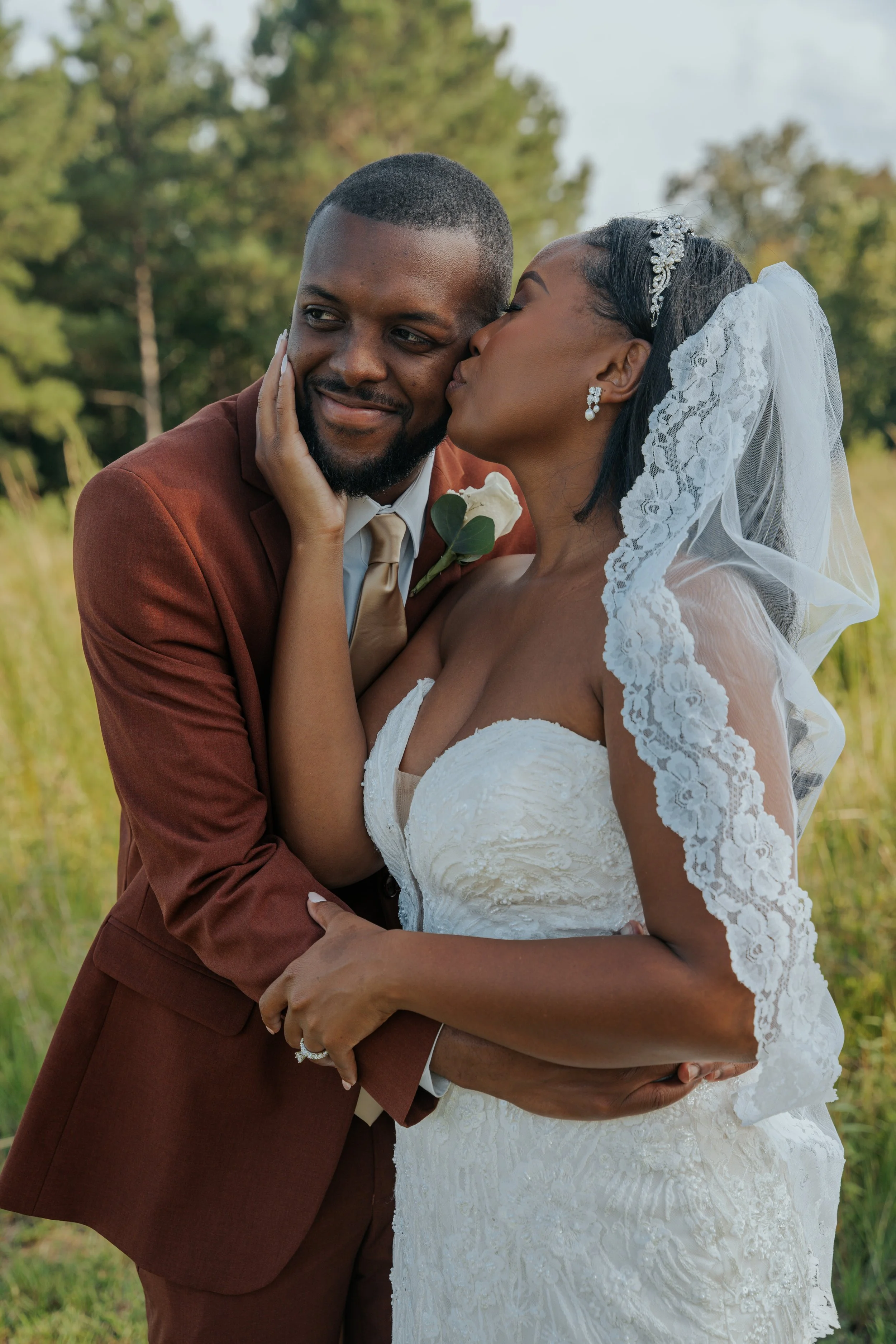 A newlywed couple embracing outdoors, the bride is giving the groom a kiss on the cheek, with a background of trees and greenery.