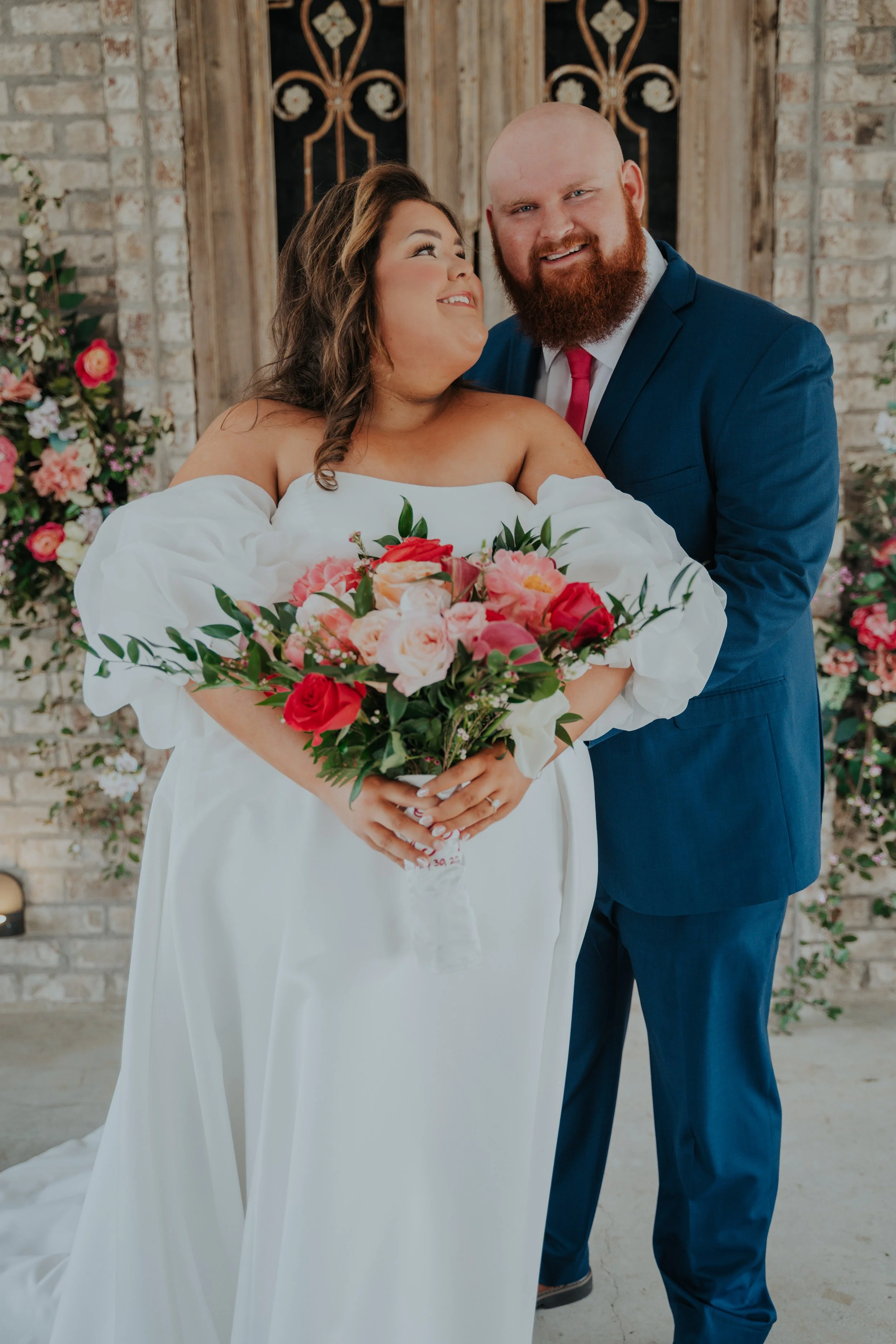 A bride holding a bouquet of pink, red, and white flowers, standing next to a groom in a blue suit, in front of a floral backdrop.