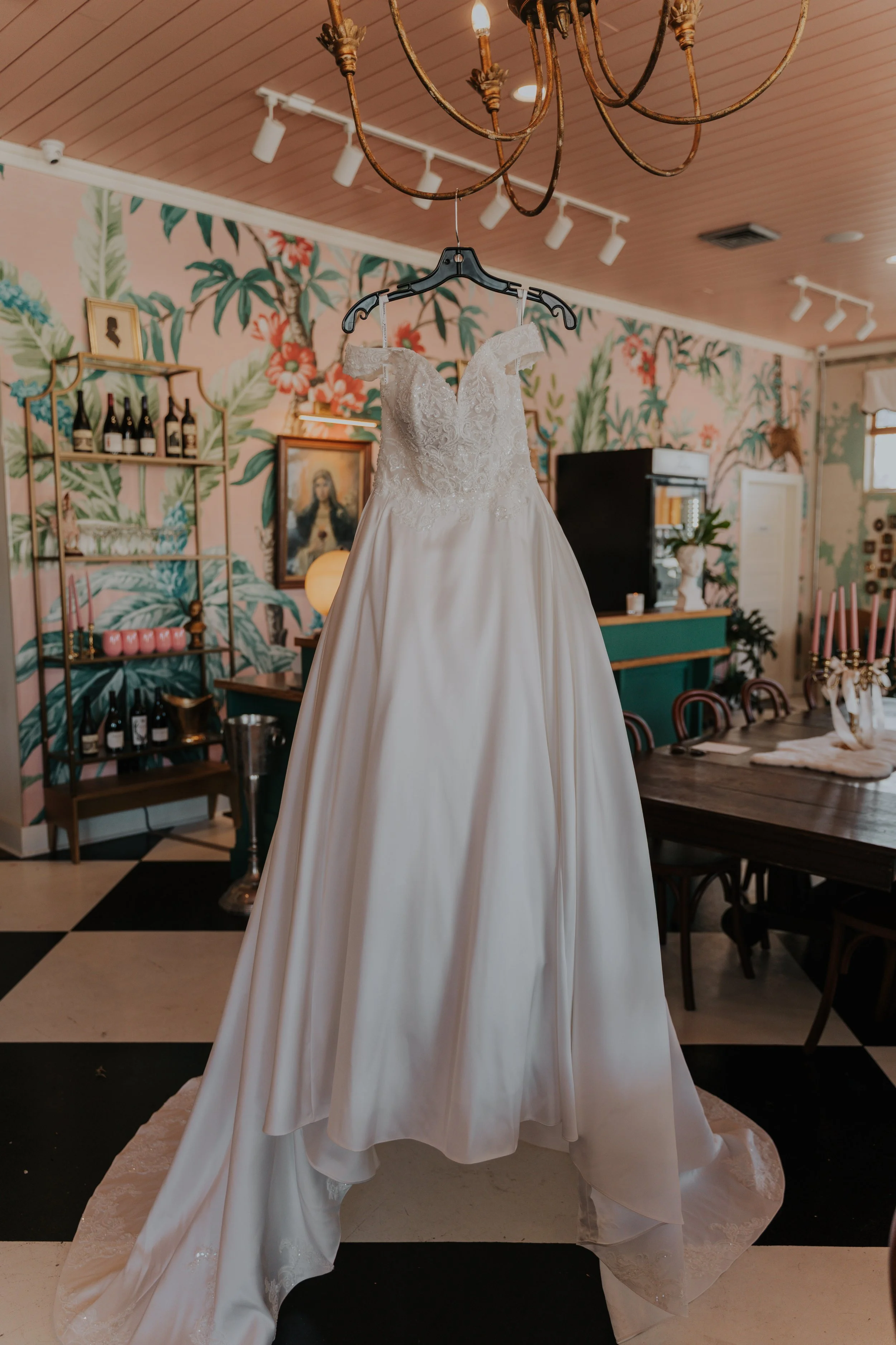 Wedding dress hanging in a room with tropical-themed wallpaper, chandelier, and dining table.