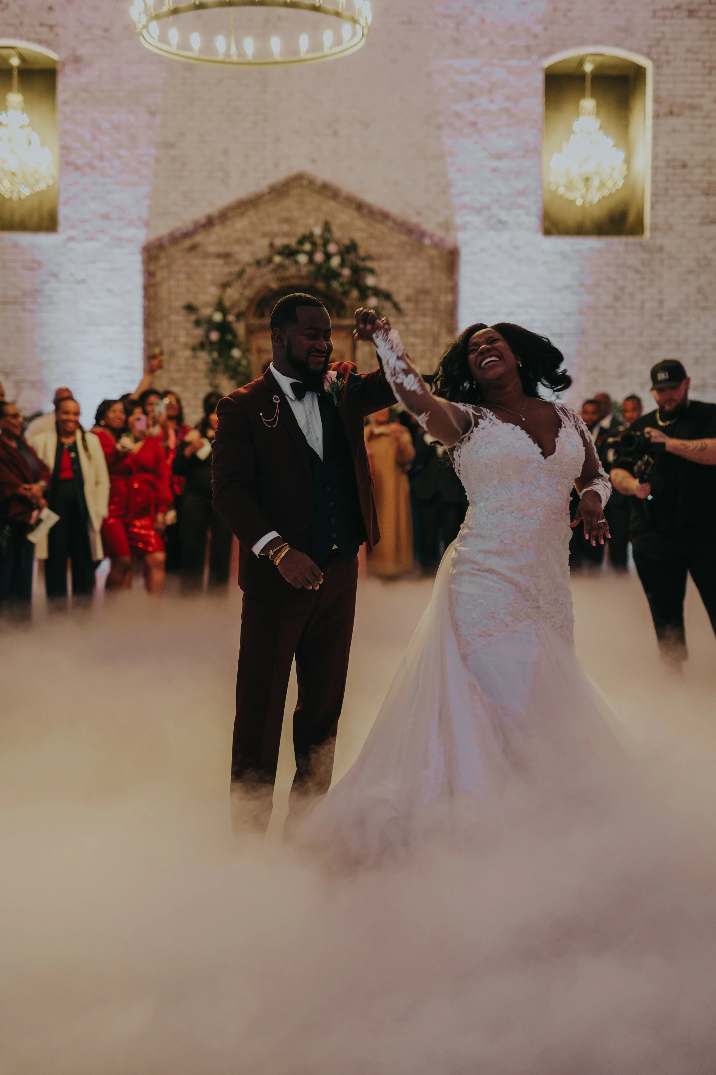 A newlywed couple dancing at their wedding reception, surrounded by guests and a fog machine, in a church with brick walls and chandeliers.