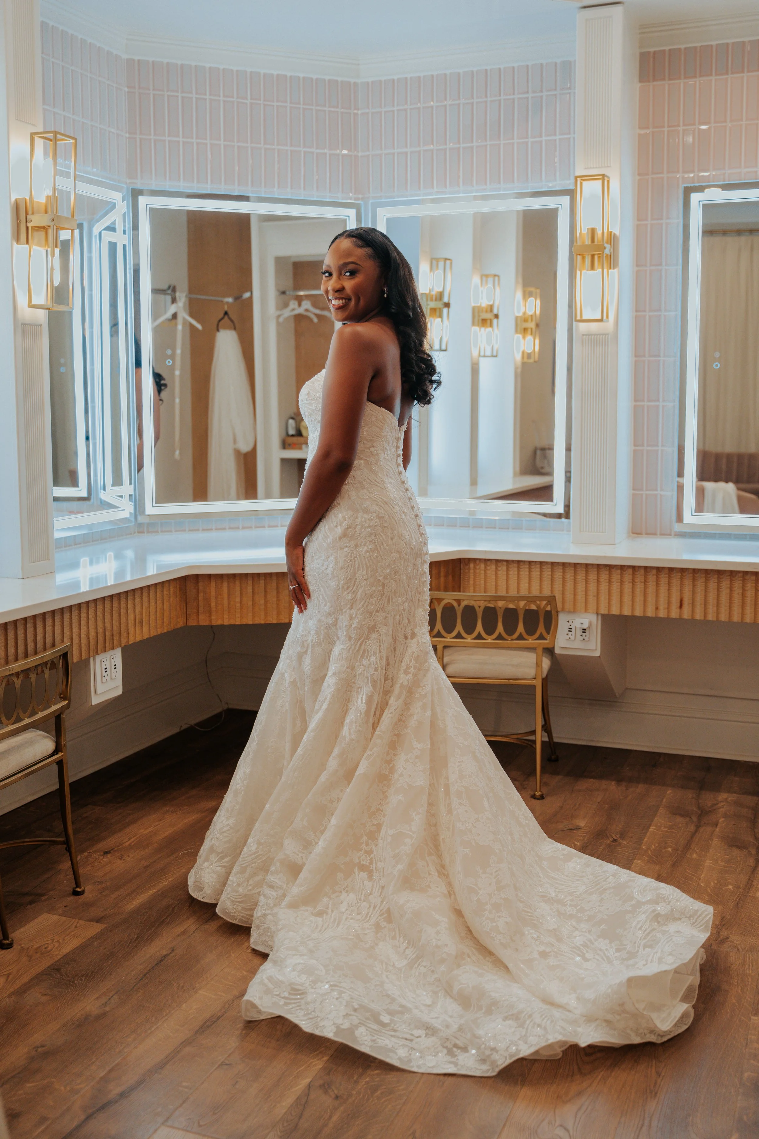 A woman in a strapless white wedding dress with a mermaid silhouette standing in front of a mirror in a dressing room with pink tiled walls and warm lighting.