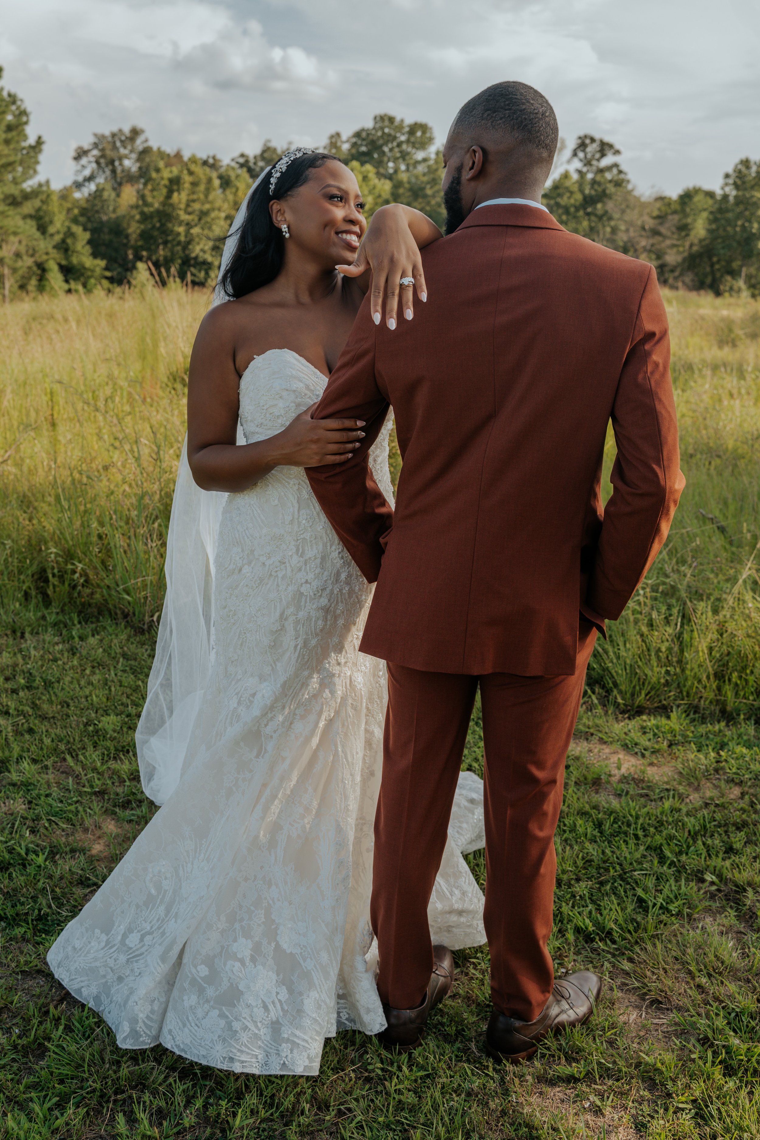 A bride in a strapless white lace wedding gown and a groom in a brown suit standing in a grassy field with trees in the background, sharing an intimate moment.