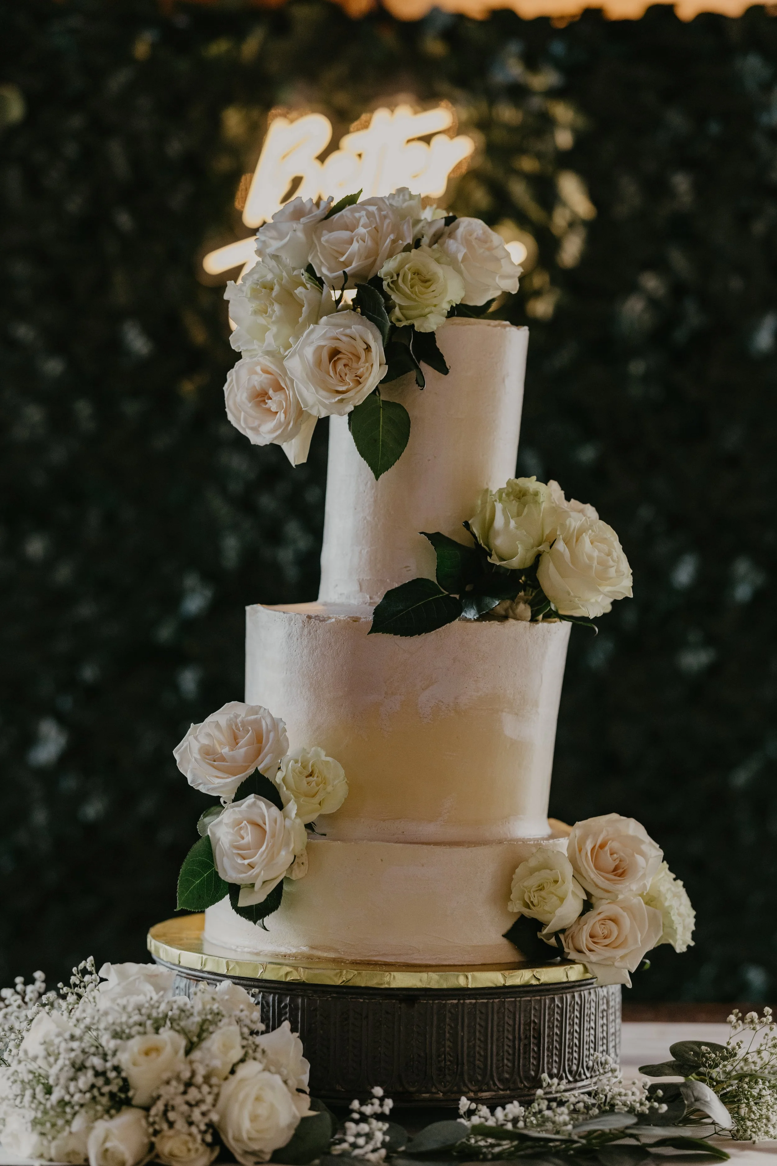 Three-tier white wedding cake decorated with white roses and greenery, with a neon sign in the background.