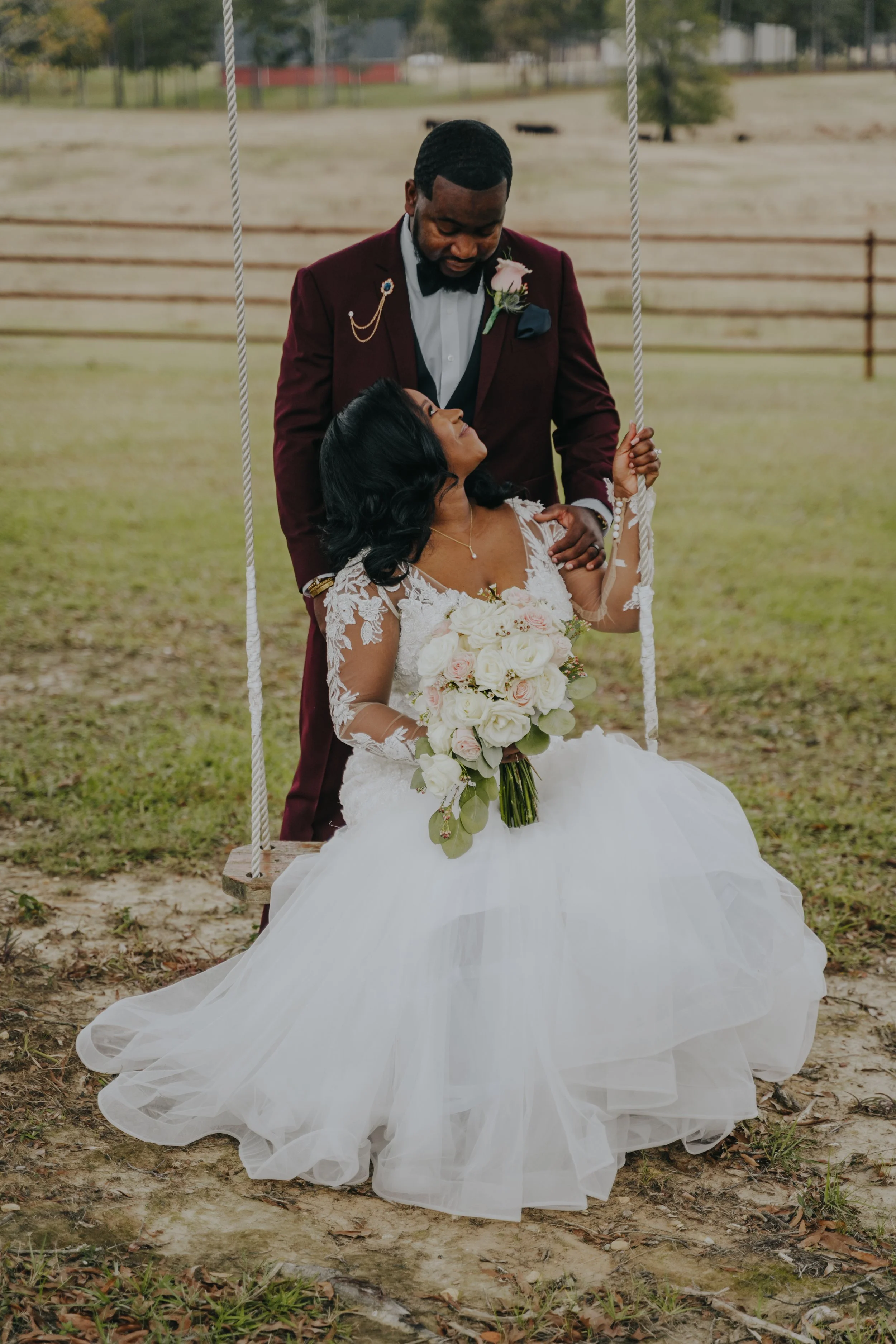 A bride sitting on a swing in a field, holding a bouquet of white and pink roses, looking up at her groom who is standing beside her, wearing a burgundy suit and black bow tie.