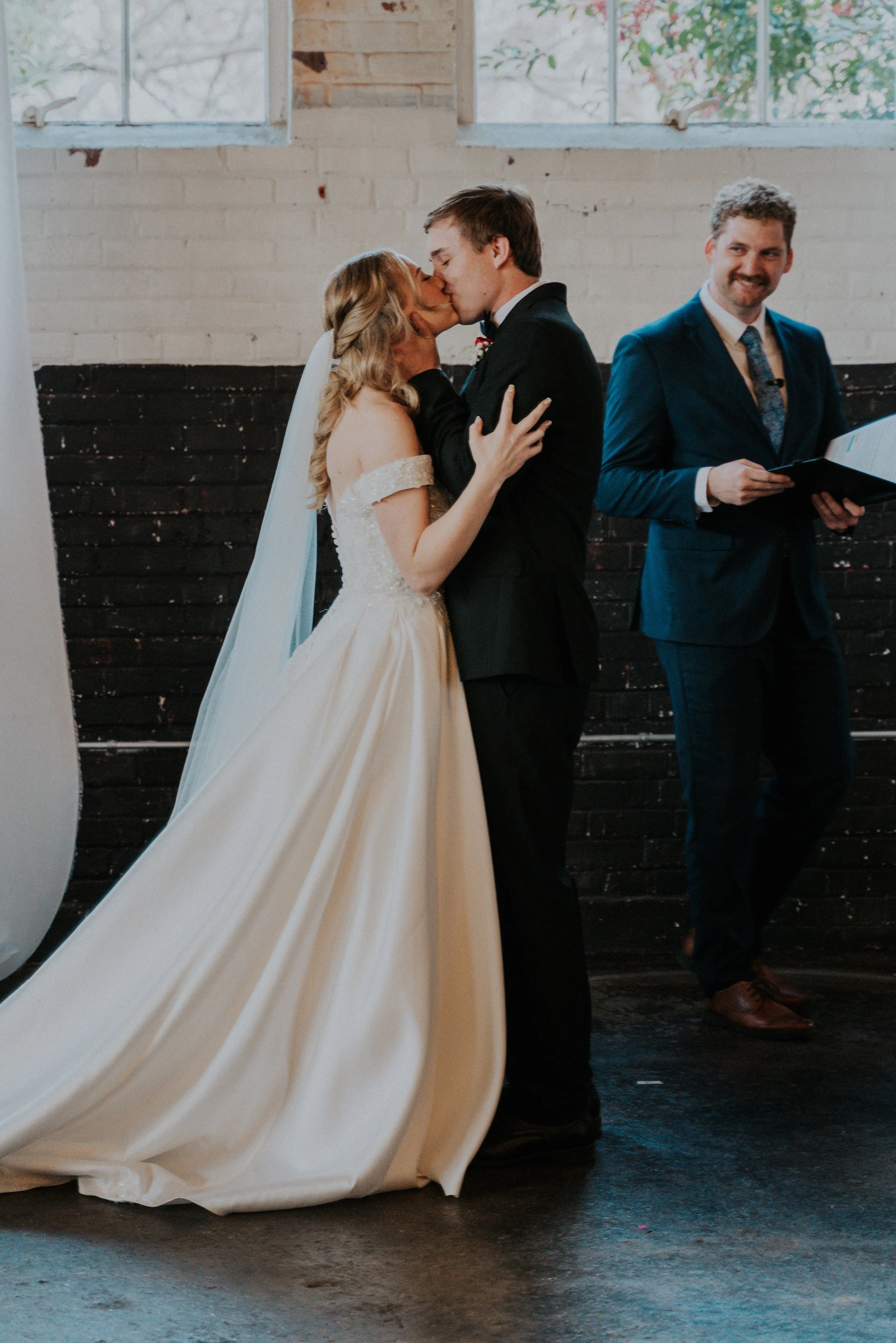A bride and groom kiss during their wedding ceremony, with an officiant standing nearby holding a booklet, inside a rustic venue with brick walls and large windows.
