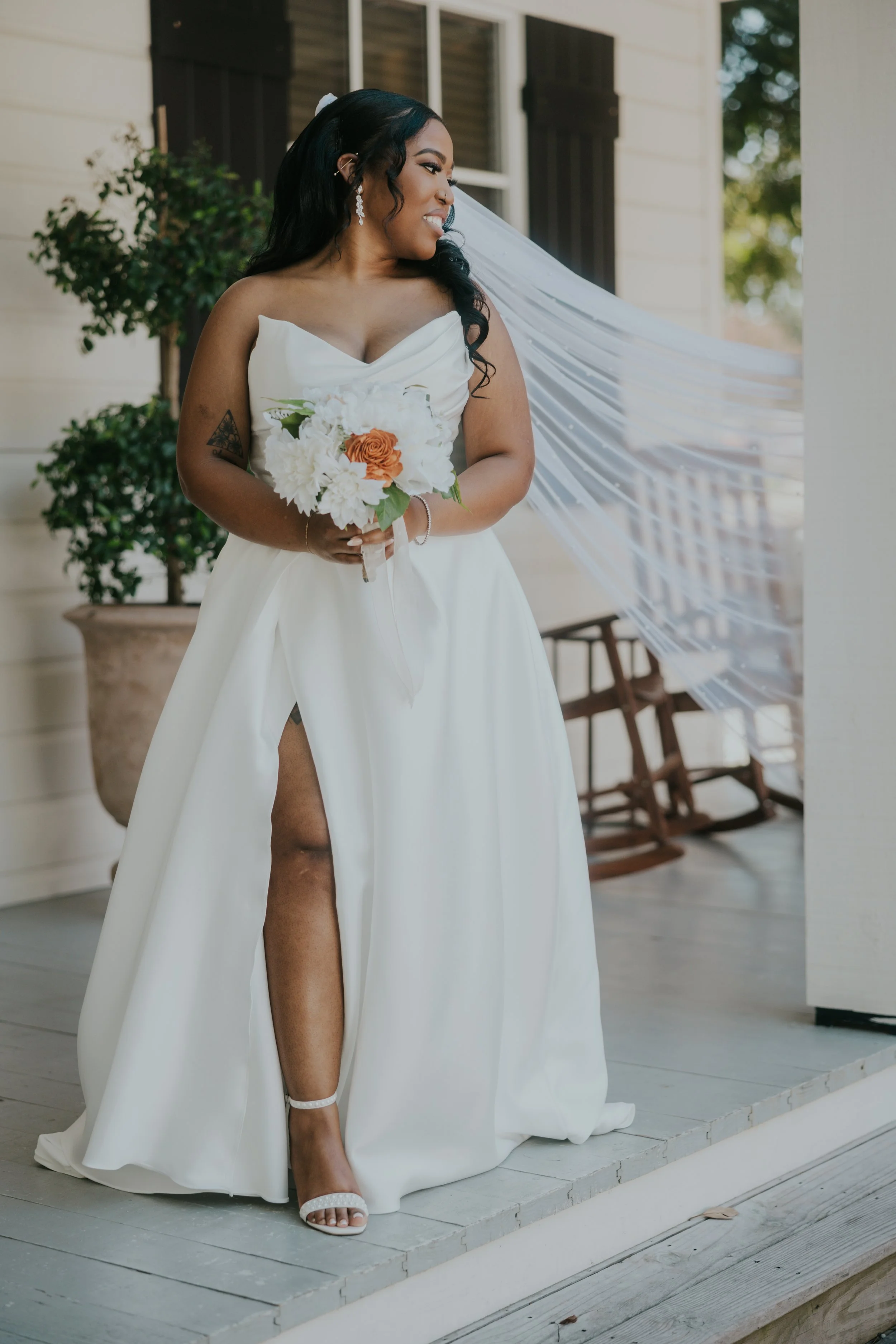 A bride standing on a porch, wearing a white wedding dress with a slit, holding a bouquet of white and orange flowers, with a veil flowing behind her, smiling and looking to her right.