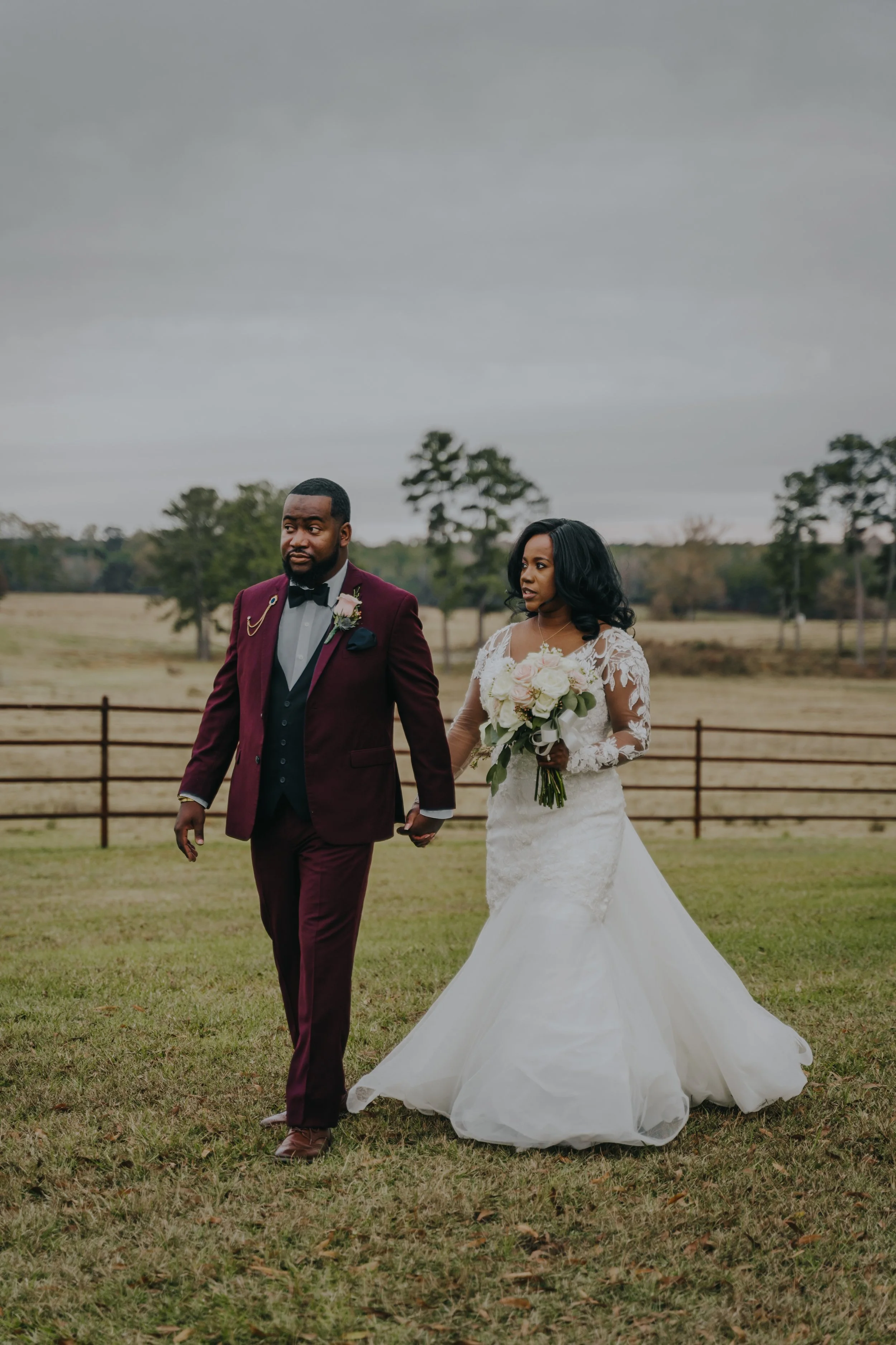 A newlywed couple holding hands and walking on a grassy field outdoors, with trees and a fence in the background, under an overcast sky.