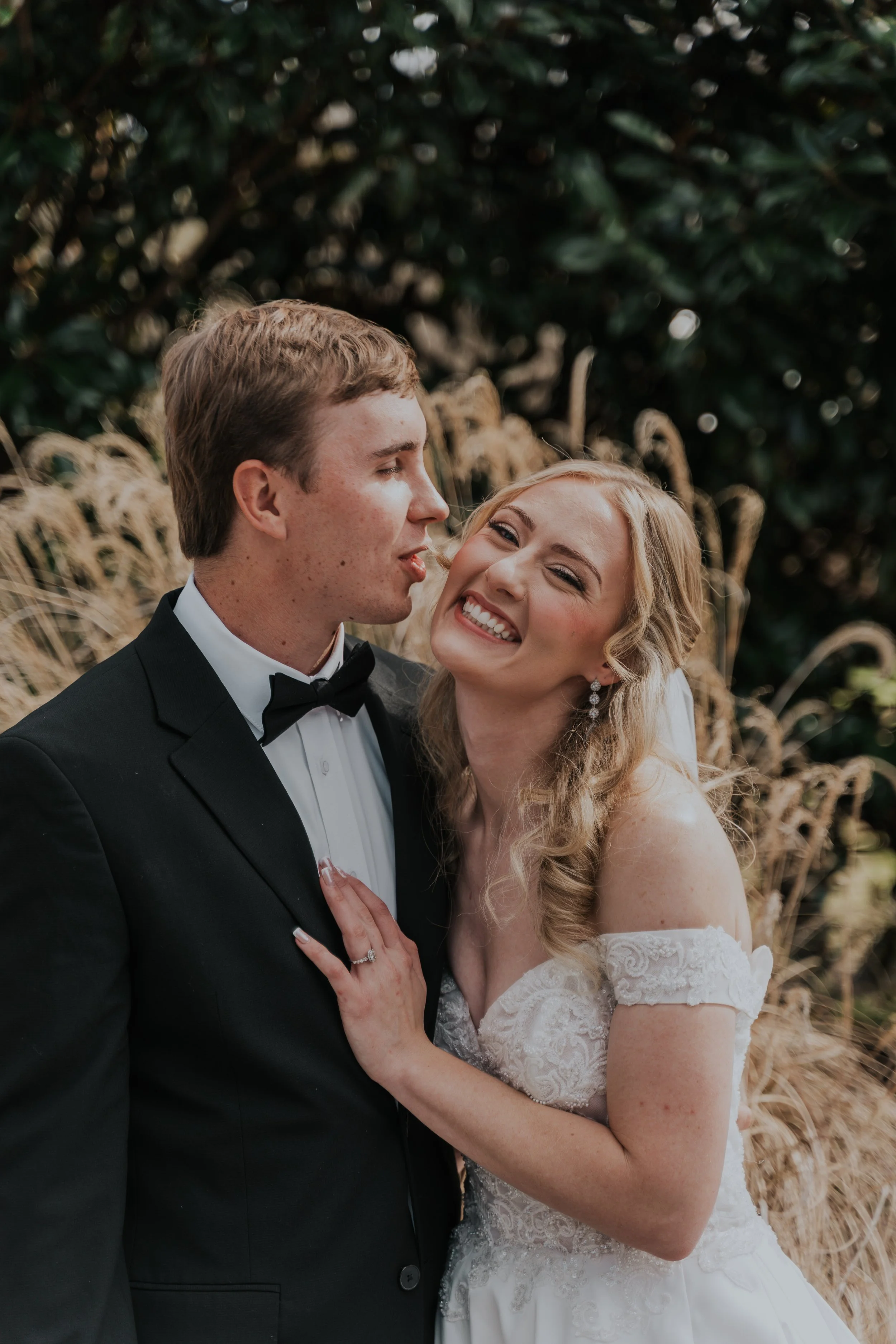 Bride and groom smiling and embracing outdoors during wedding, with natural greenery and dried plants in background.