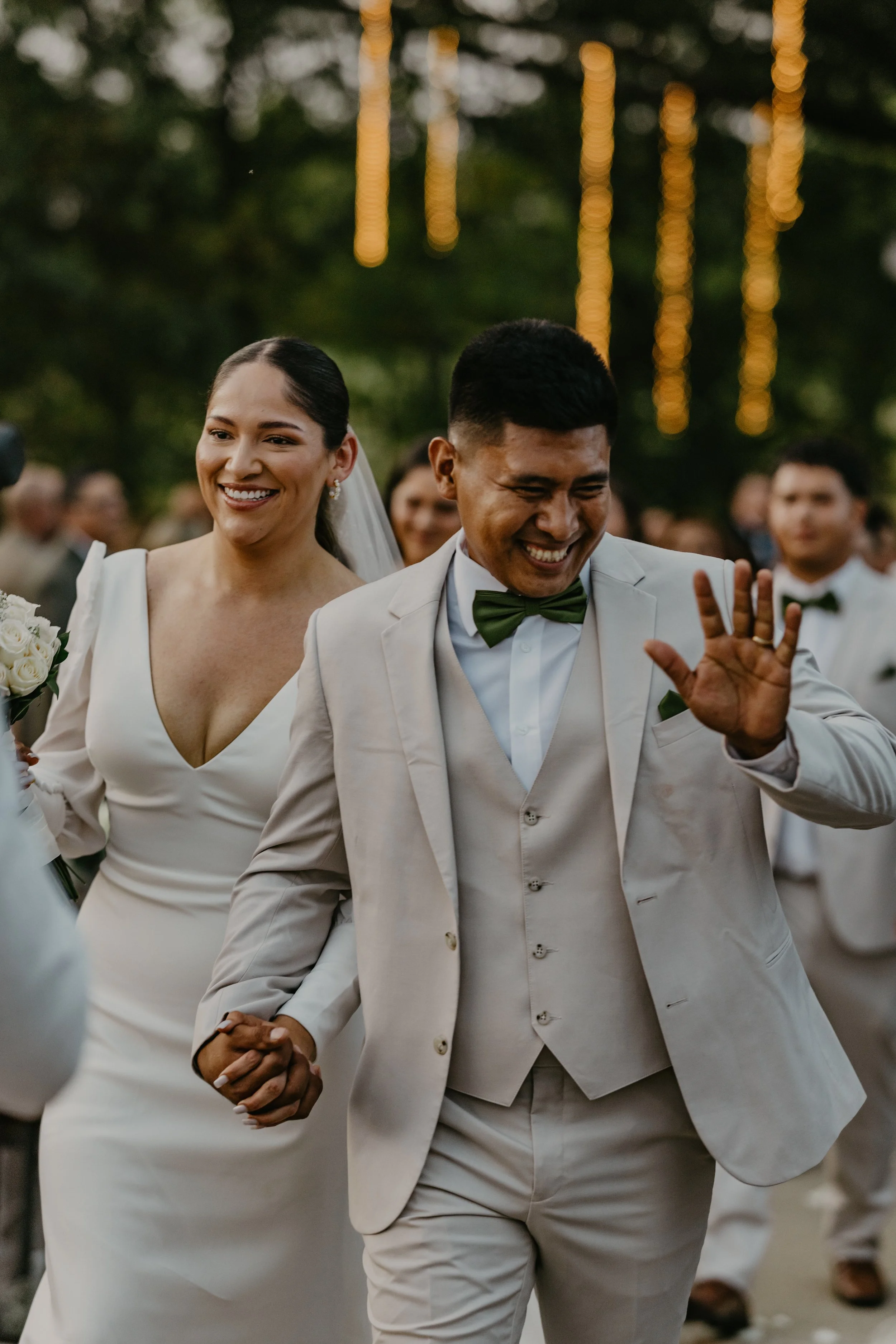 A bride and groom holding hands and smiling during their outdoor wedding celebration, with guests and string lights in the background.