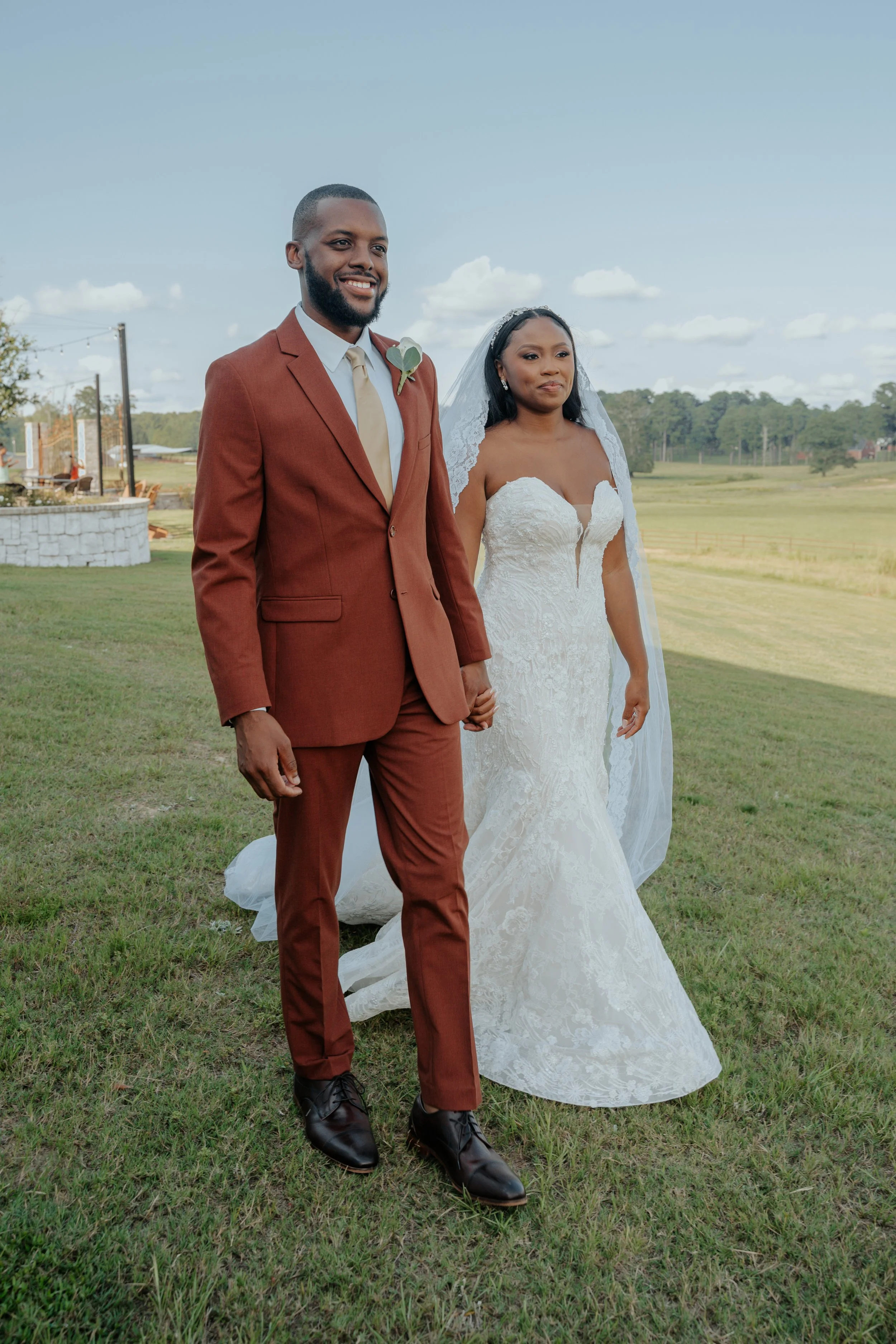 A newlywed couple holding hands outdoors on a grassy field, with the bride in a white lace wedding gown and veil, and the groom in a brown suit and cream tie, smiling as they pose for a photo.