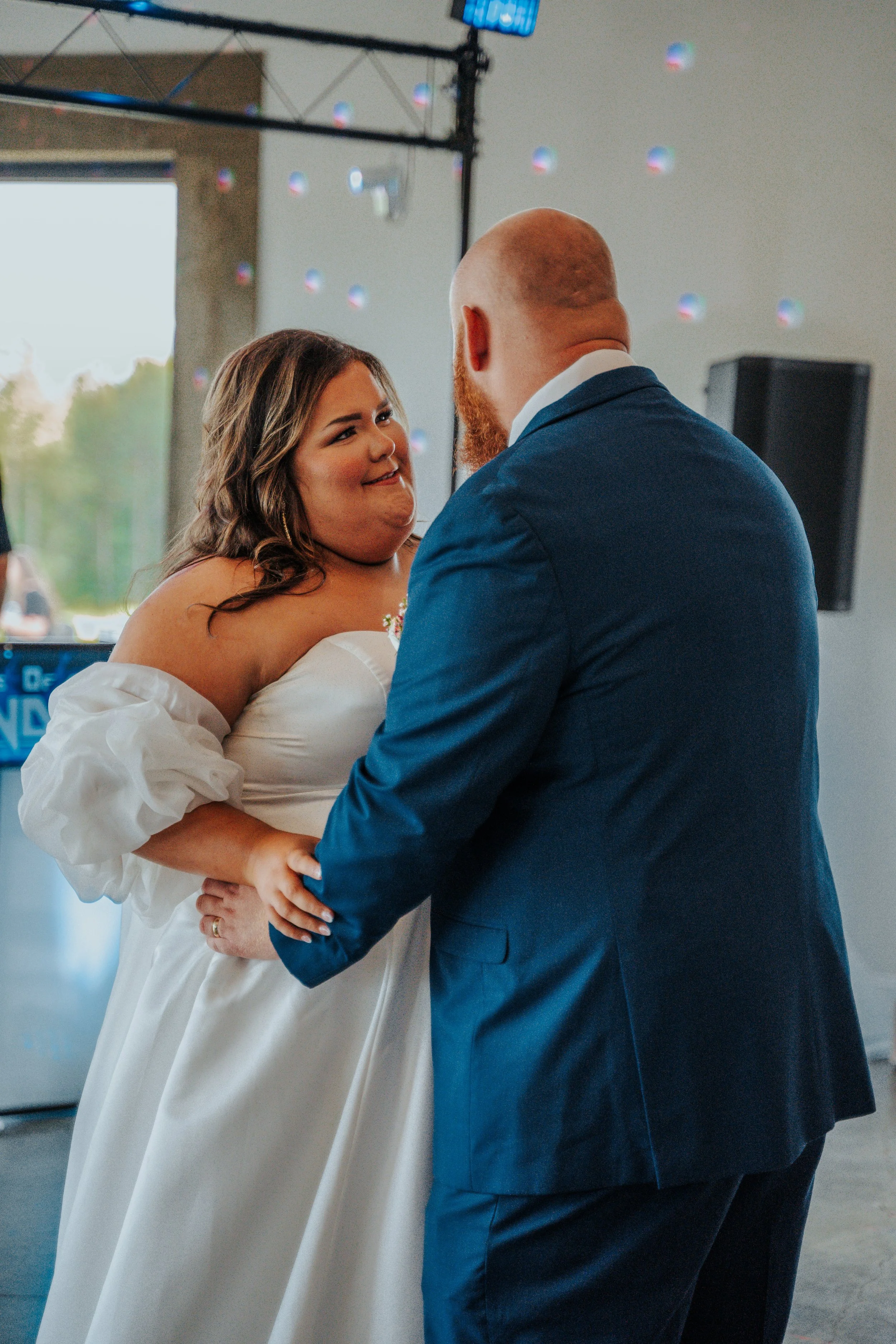 A bride and groom dancing at their wedding reception, looking into each other's eyes, indoors with a large window and some disco lights in the background.
