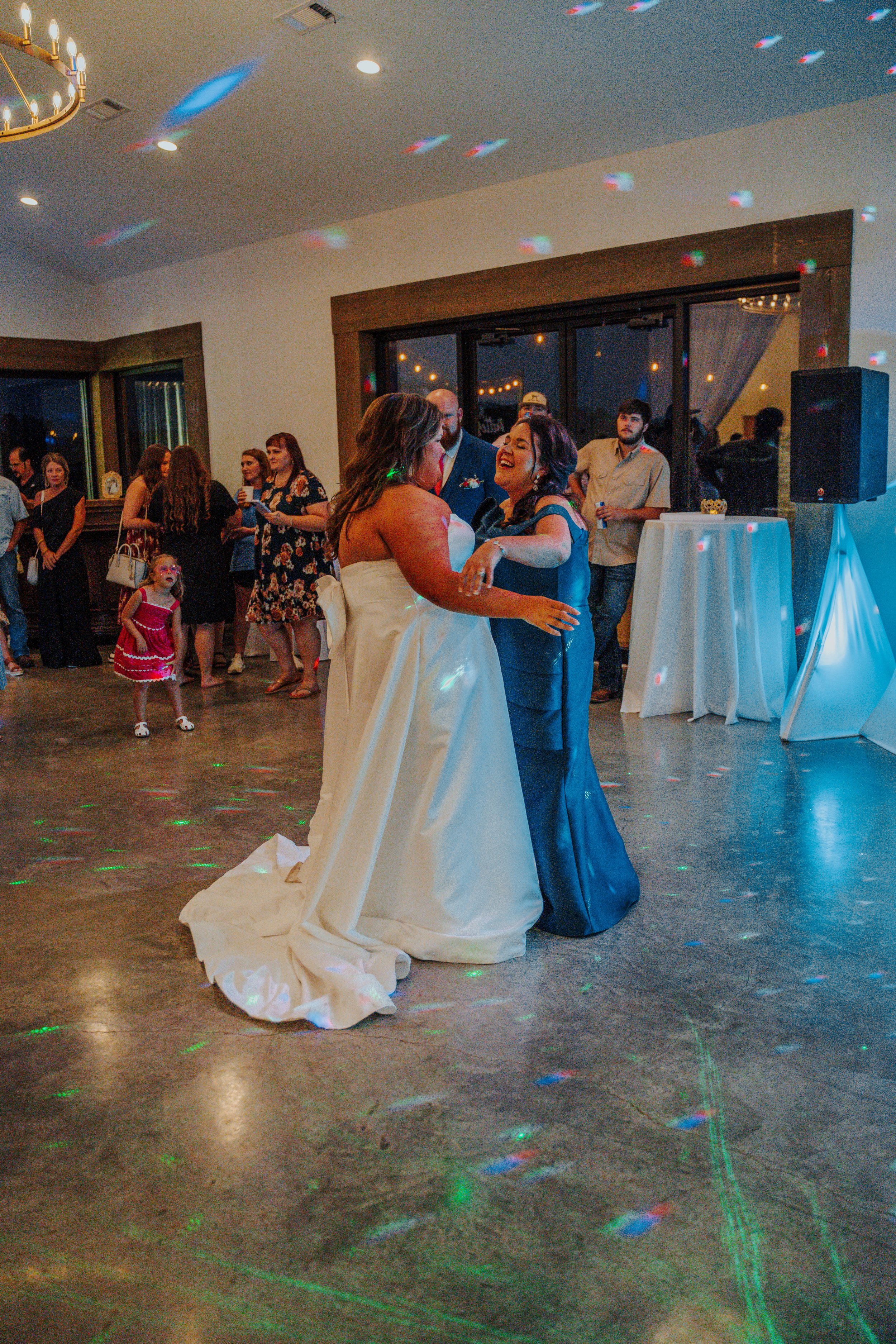 Two women dancing at a celebration, with one wearing a wedding dress and the other wearing a dark blue dress, in a decorated event space with guests watching and colorful lights on the ceiling.