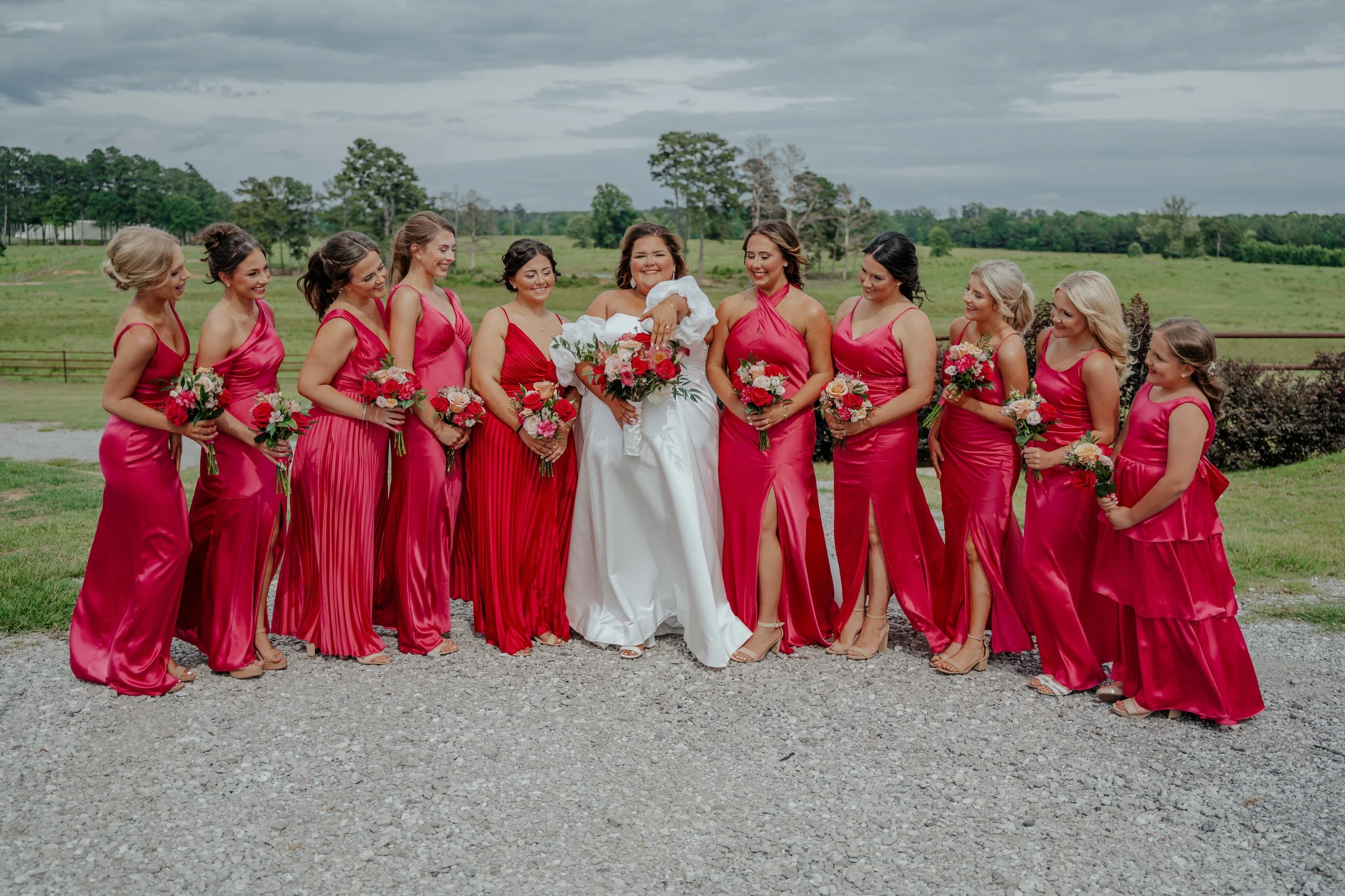 Group of women in pink bridesmaid dresses and a bride in a white gown standing outdoors on a gravel surface with green fields and cloudy sky in the background, holding bouquets of pink and white flowers, smiling and looking at each other.