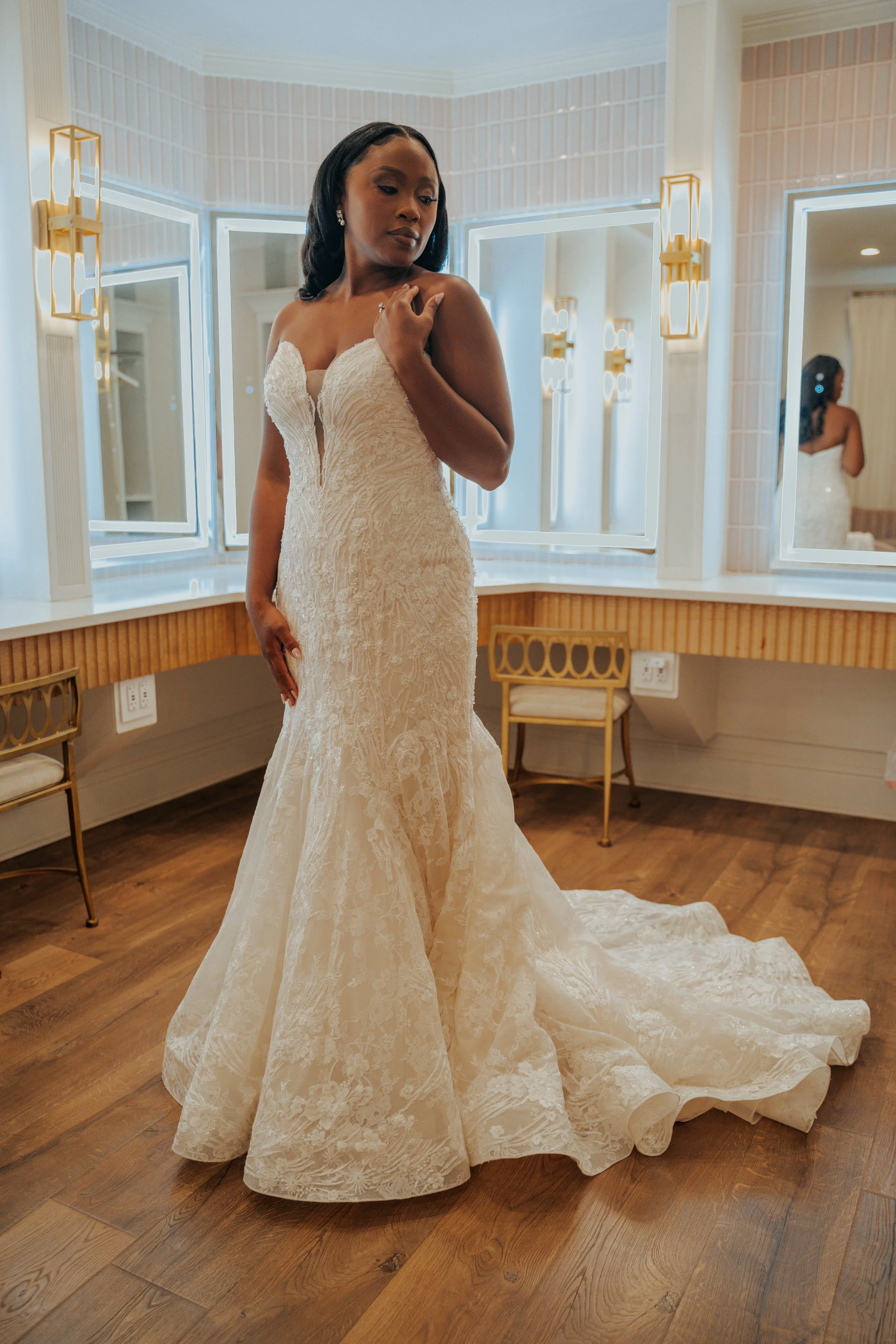 A woman in a white lace wedding dress standing in front of mirrors in a well-lit room.