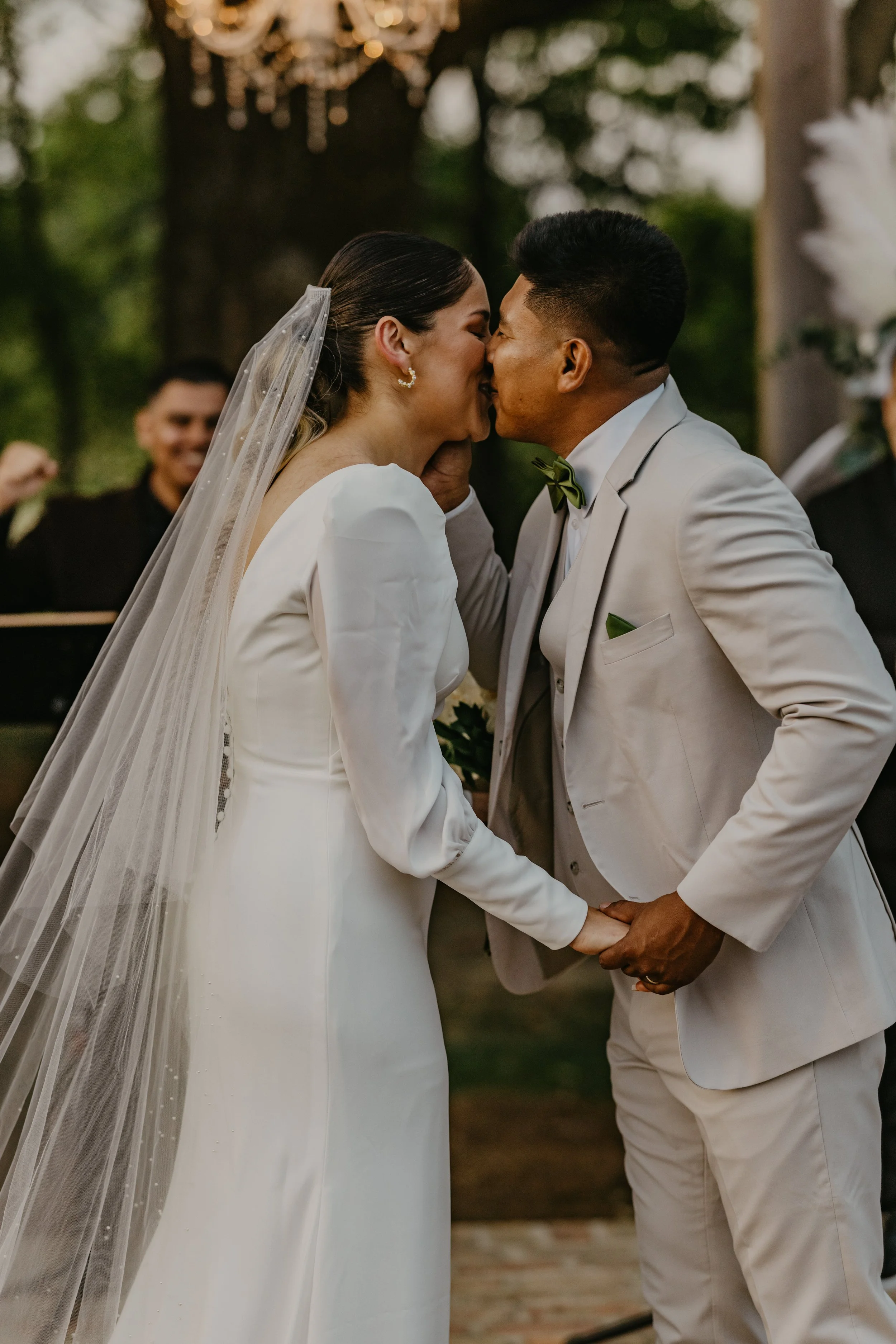 A bride and groom share a kiss during their wedding ceremony outdoors, holding hands, with a pianist and greenery in the background.