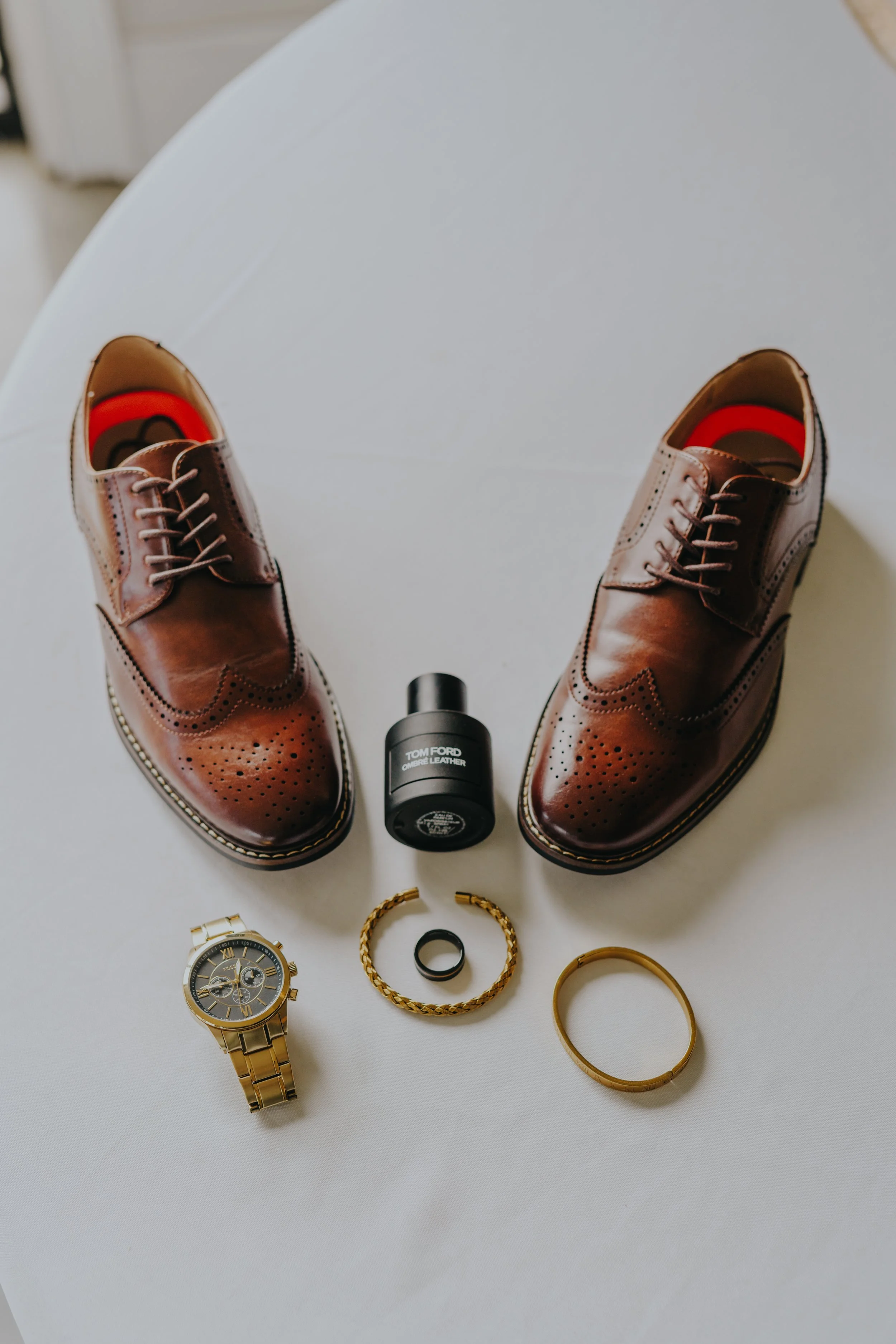 Pair of brown leather dress shoes with decorative broguing, gold watch, black ring, gold bracelet, gold bangle, fragrance bottle, and white surface background.