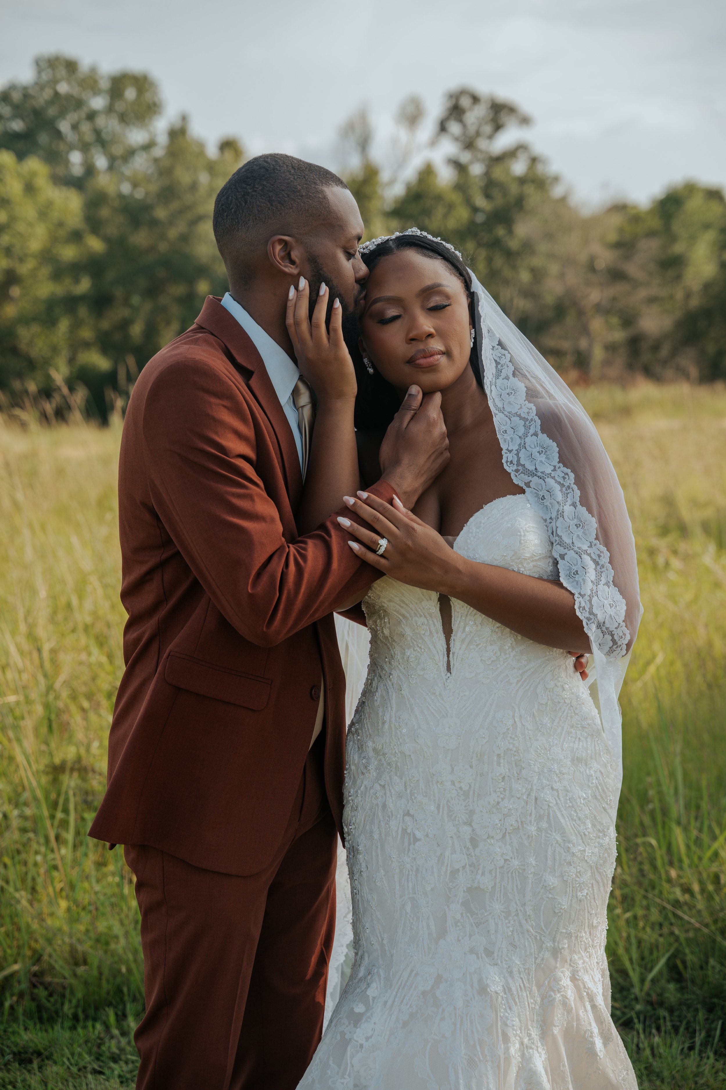 A bride and groom share a tender moment in a grassy outdoor setting, with trees in the background. The groom, wearing a brown suit, gently holds the bride's face as she closes her eyes. The bride is dressed in a white lace wedding gown with a veil.