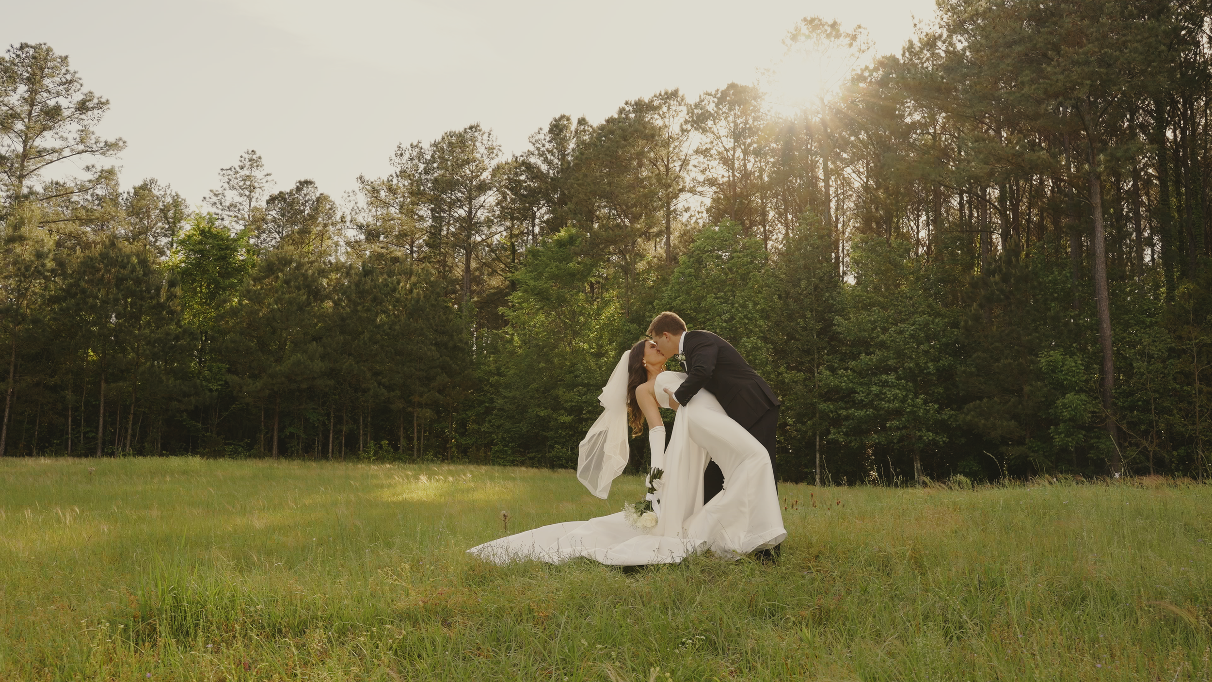 A bride and groom sharing a kiss in a grassy field with trees in the background, during sunset.
