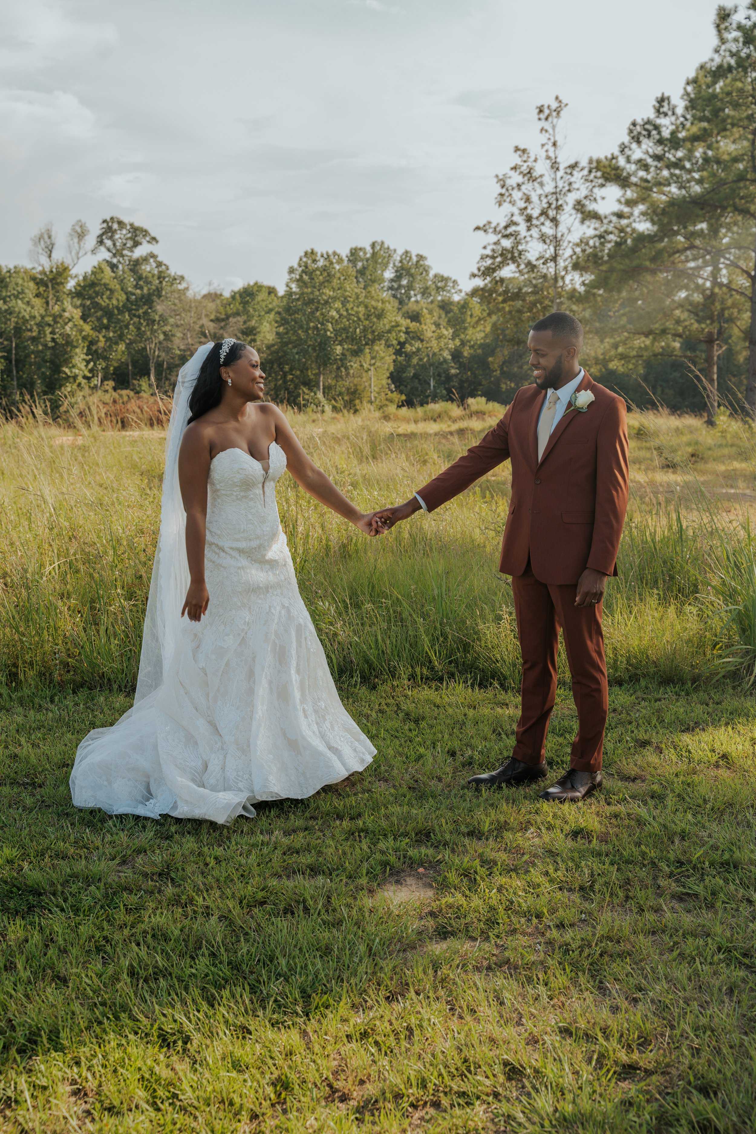 A bride and groom holding hands outdoors in a grassy field, with trees and a cloudy sky in the background.