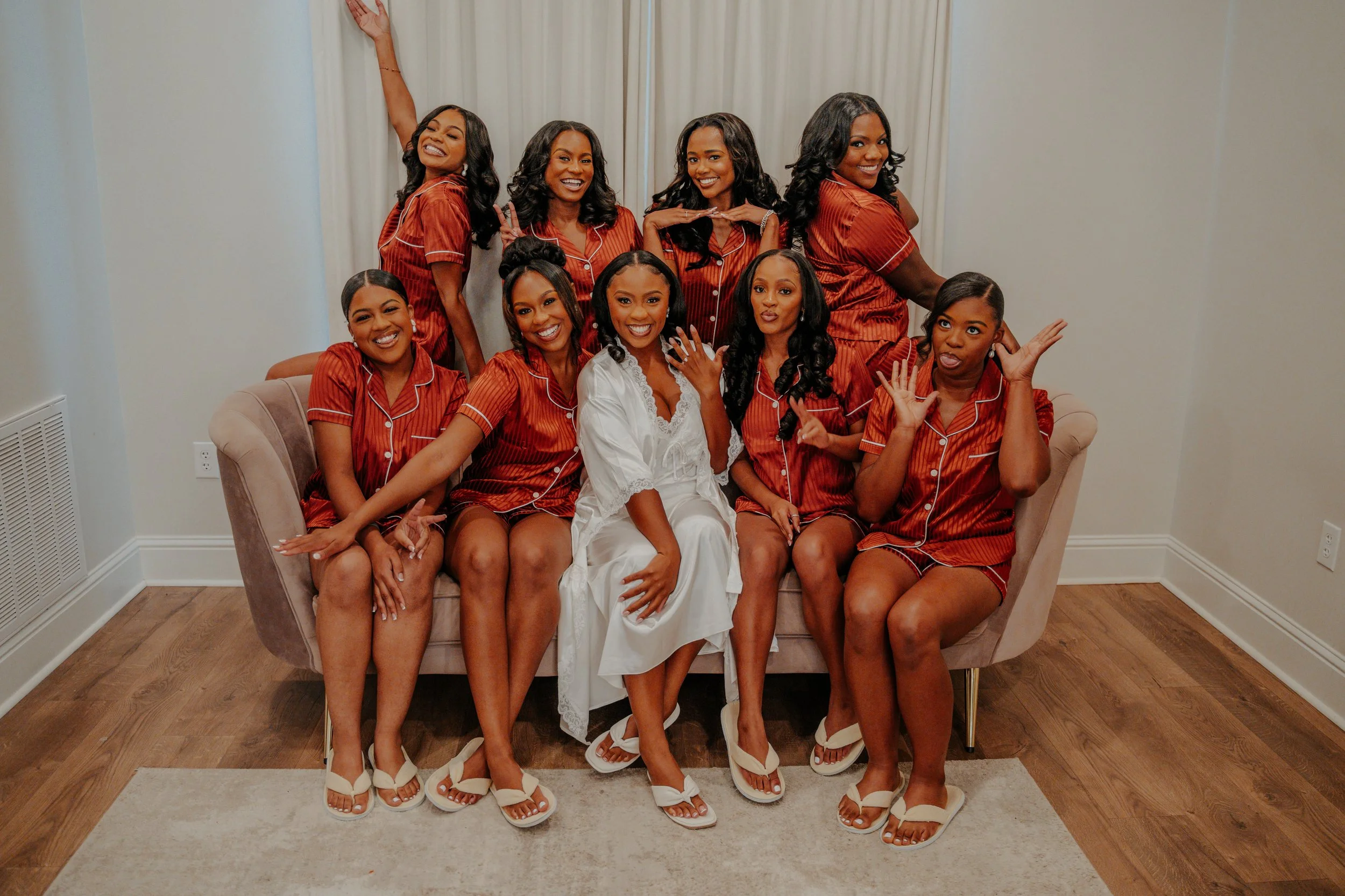 A bride in white satin robe sitting on a beige sofa surrounded by nine bridesmaids in matching red pajamas, posing playfully in a room with wooden flooring and white walls.