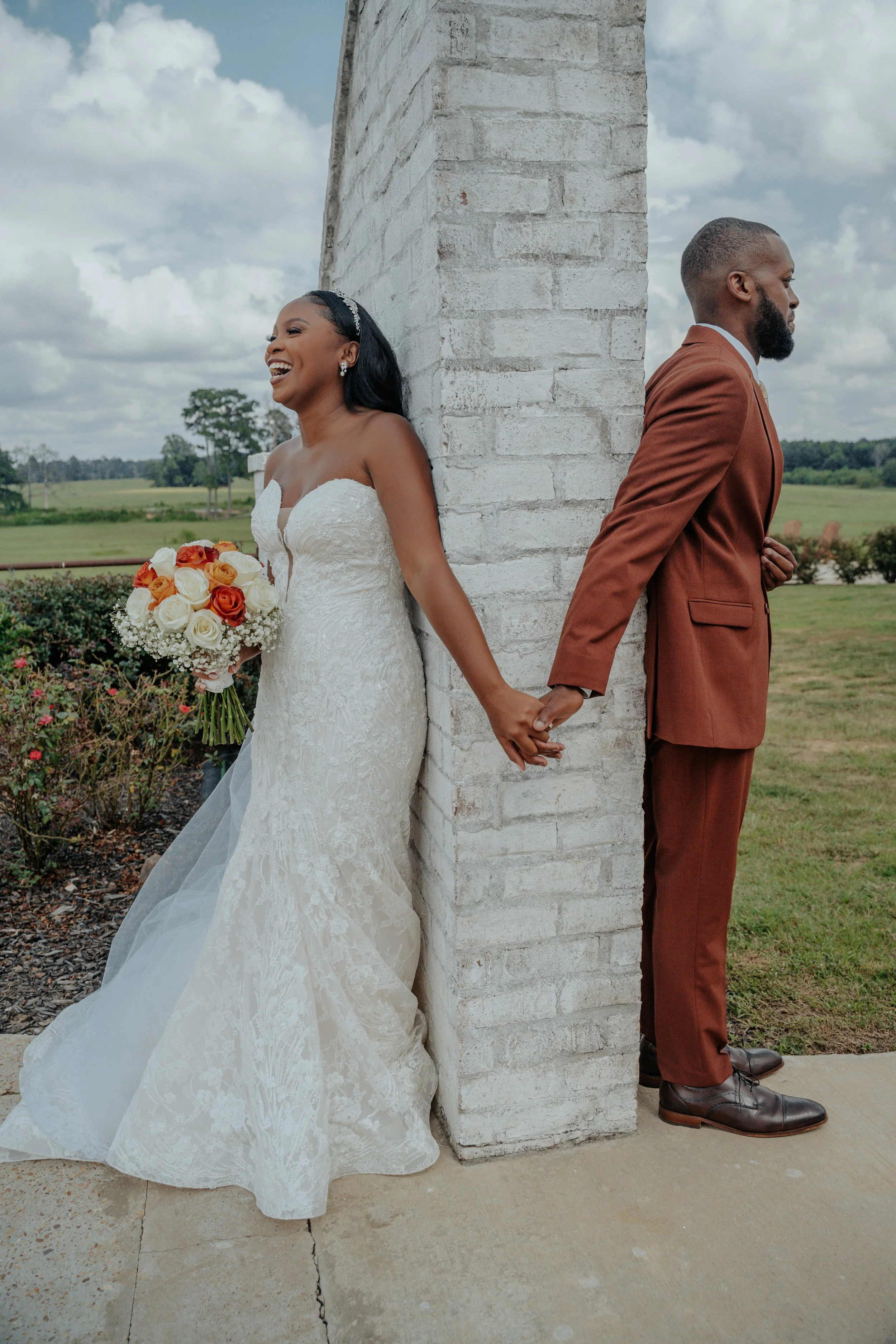 A bride and groom are standing back-to-back, holding hands behind a brick wall at an outdoor wedding venue, with open field and cloudy sky in the background.