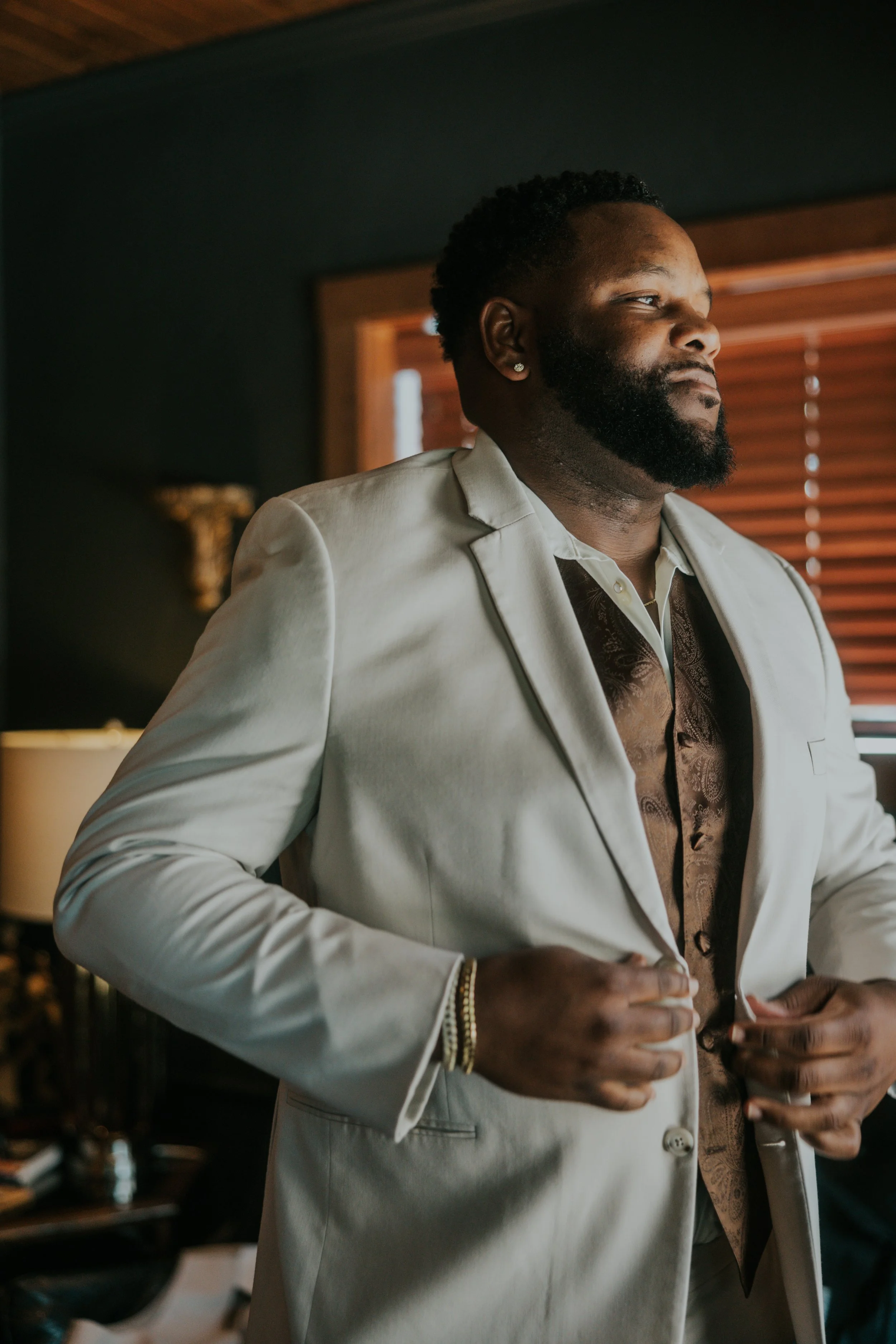 Man in cream-colored suit adjusting his suit jacket in a room with wooden blinds and a dark wall.
