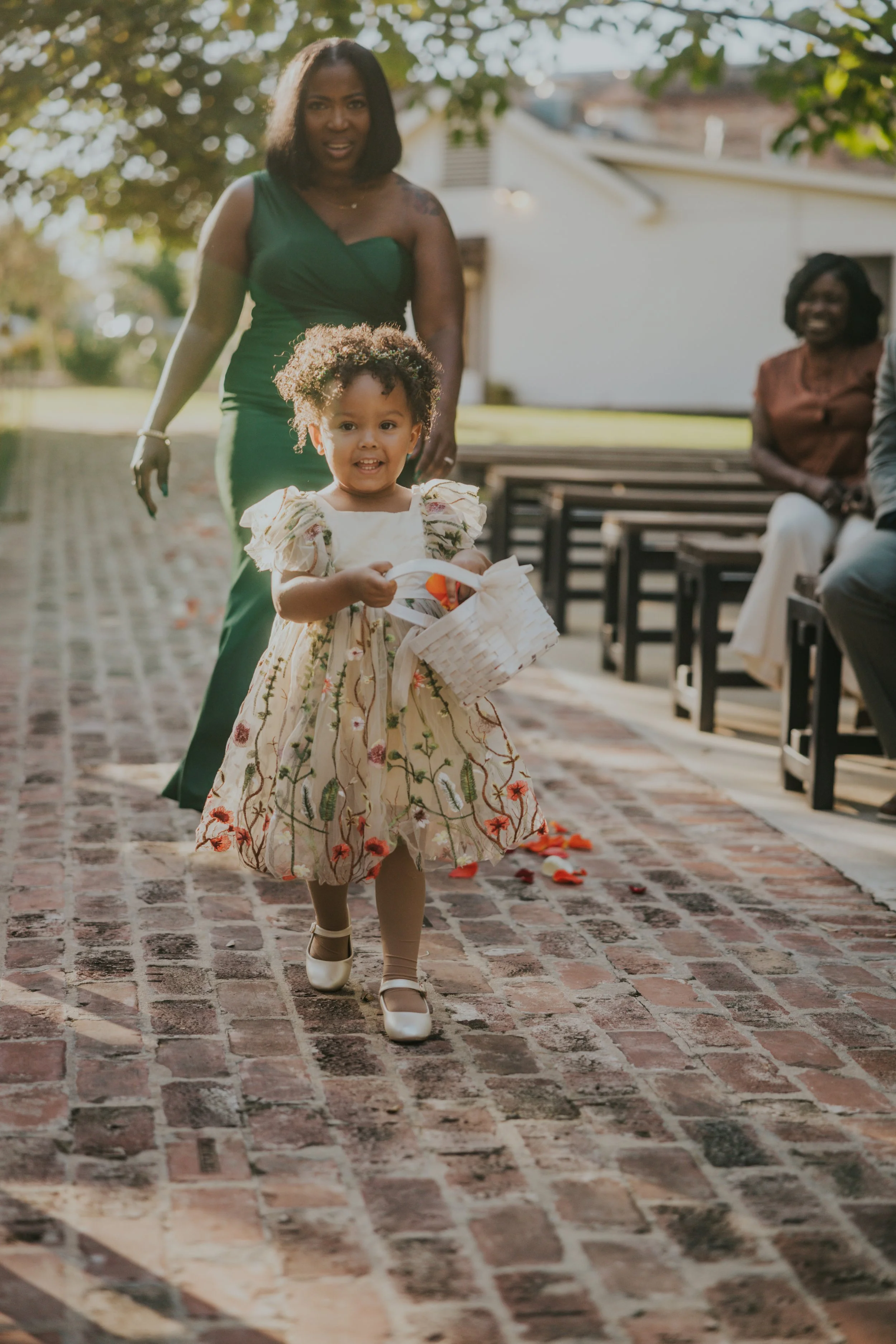 Young girl holding a small white basket walking on brick pathway outdoors, with a woman in a green dress following behind, and two women seated on benches in the background, sunlight filtering through trees.