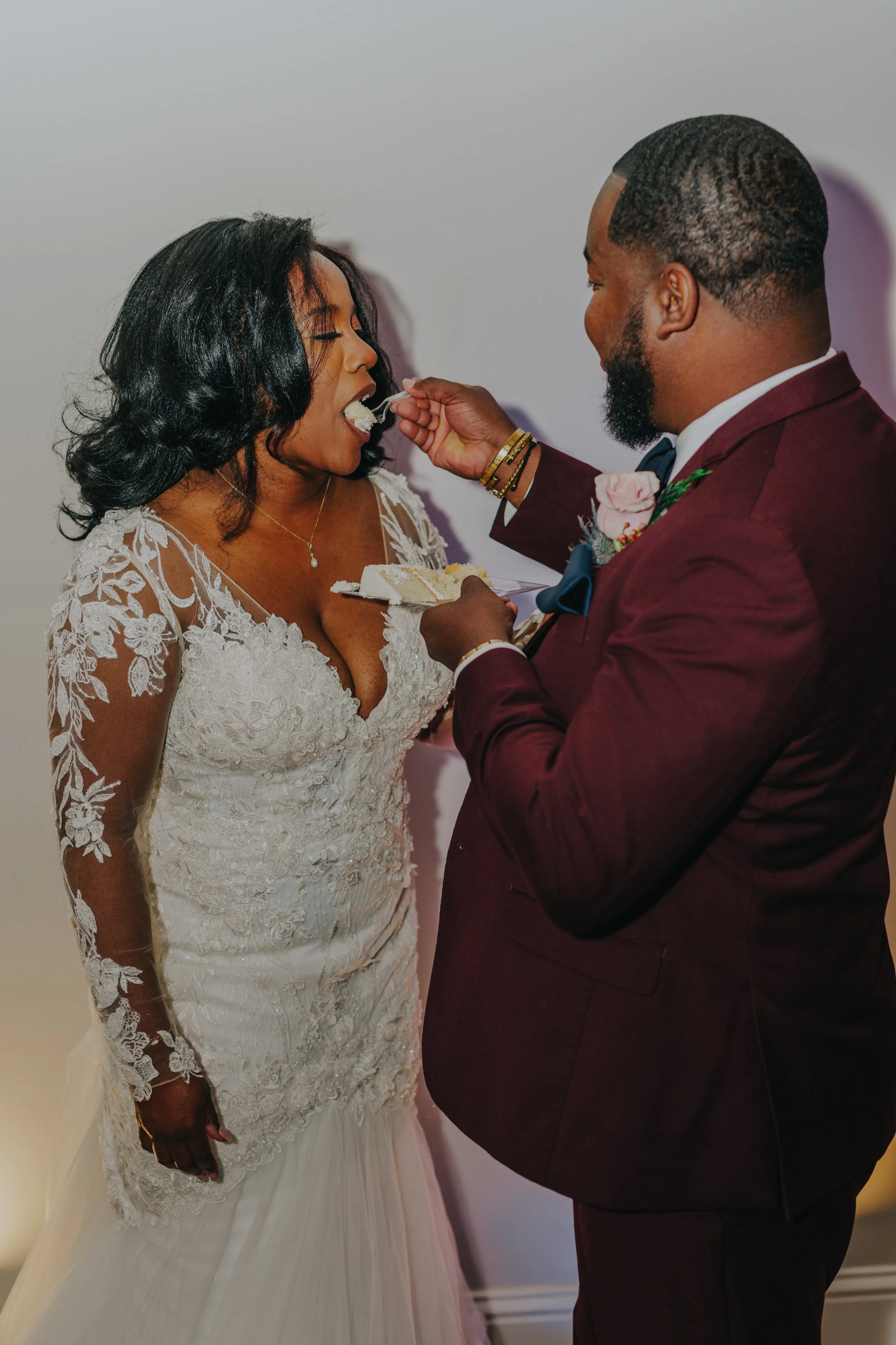 Bride in a lace wedding gown being fed cake by groom in a maroon suit.