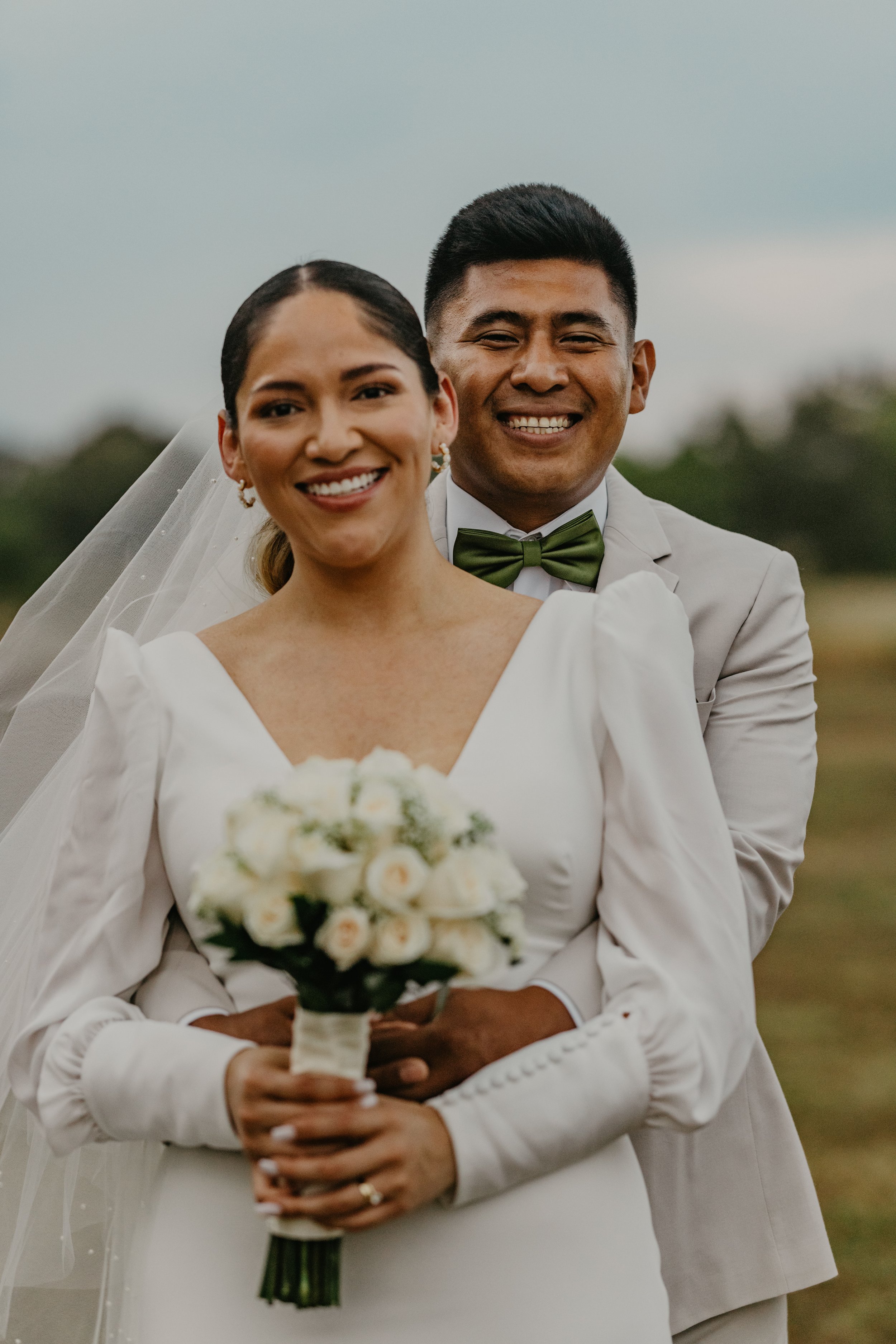 A happy bride and groom smiling outdoors on their wedding day, with the bride holding a bouquet of white roses and the groom wearing a light suit and green bow tie.