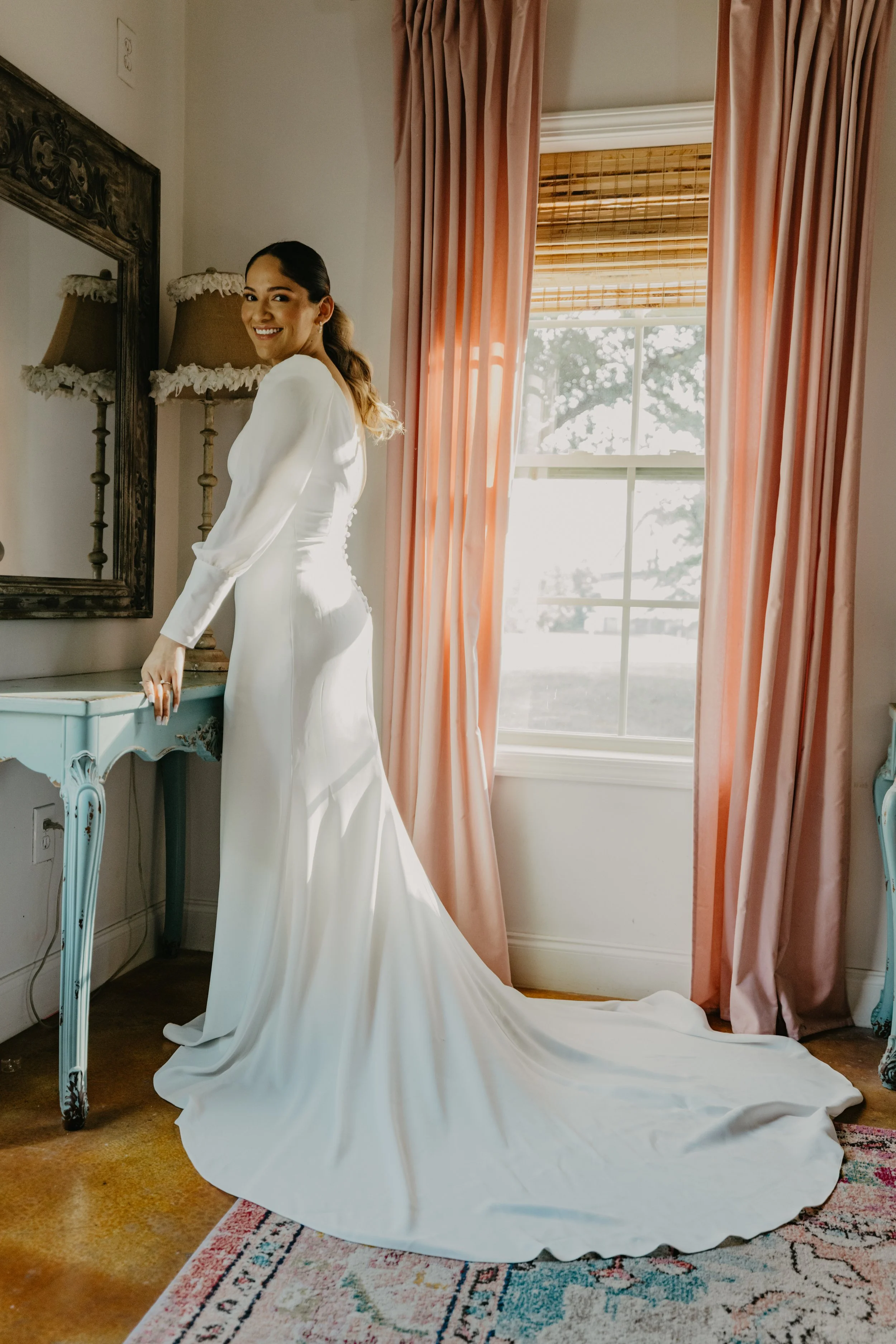 A woman in a white wedding dress standing by a window with pink curtains, smiling at the camera in a well-lit room.