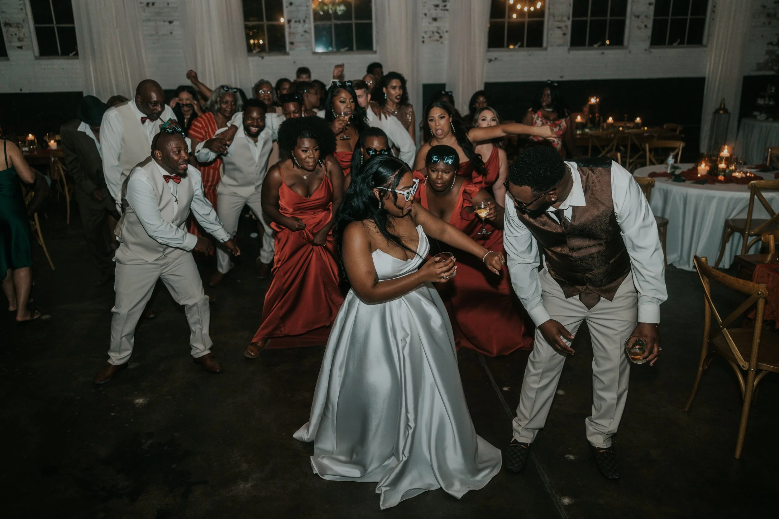People dancing at a wedding reception, with the bride in a white gown and the groom in a vest, surrounded by guests in formal attire.