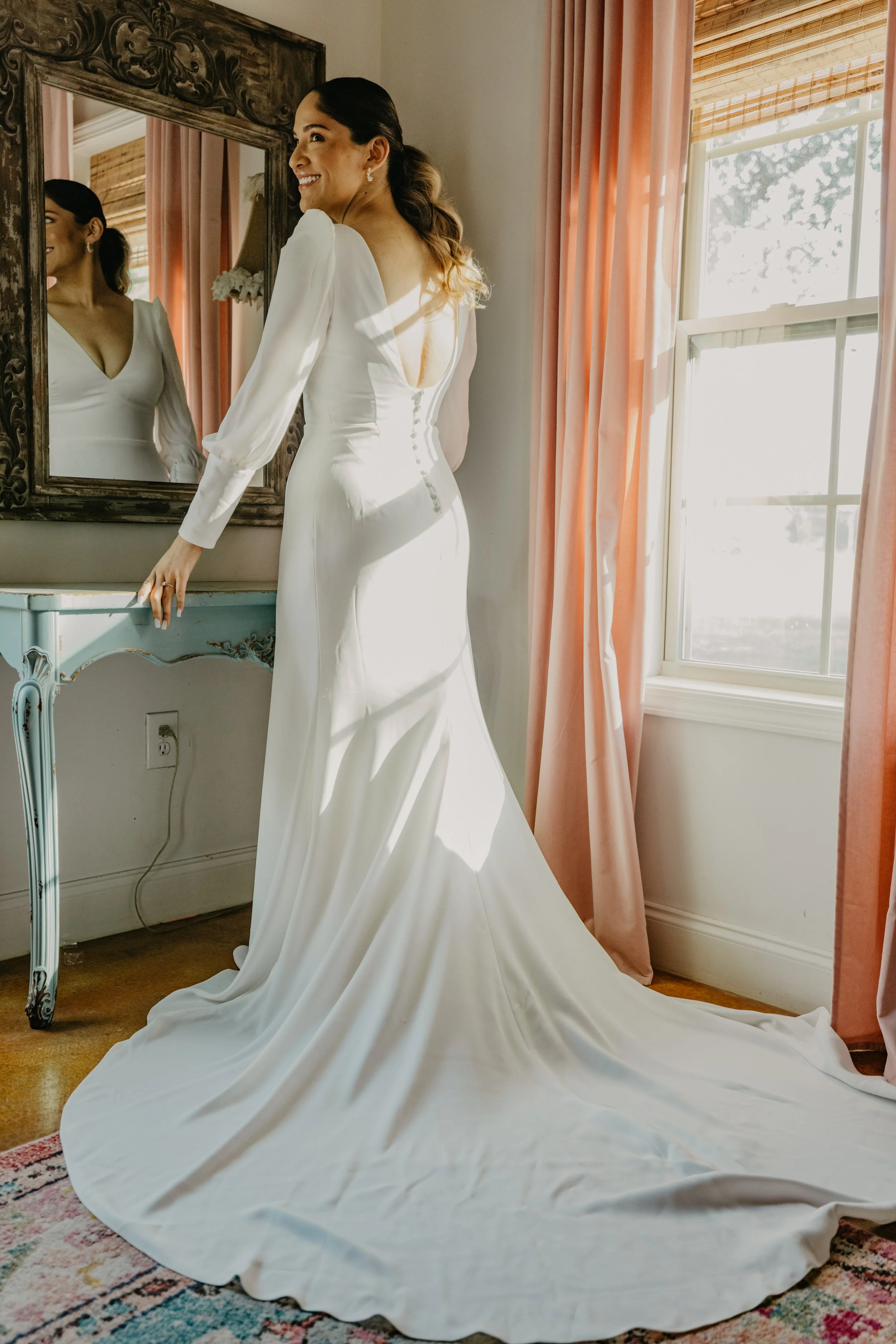 A woman in a white wedding dress standing in front of a mirror, smiling, with pink curtains and a window in the background.
