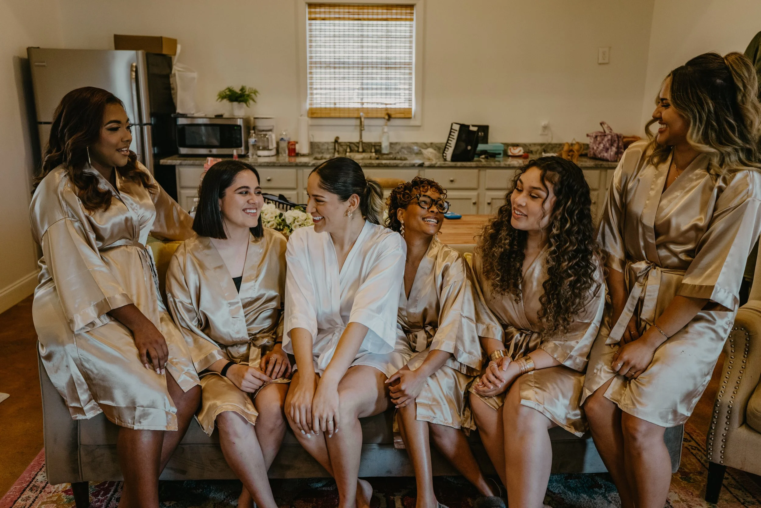 A group of seven women wearing matching satin robes sitting and standing in a cozy kitchen, smiling, and sharing a happy moment.