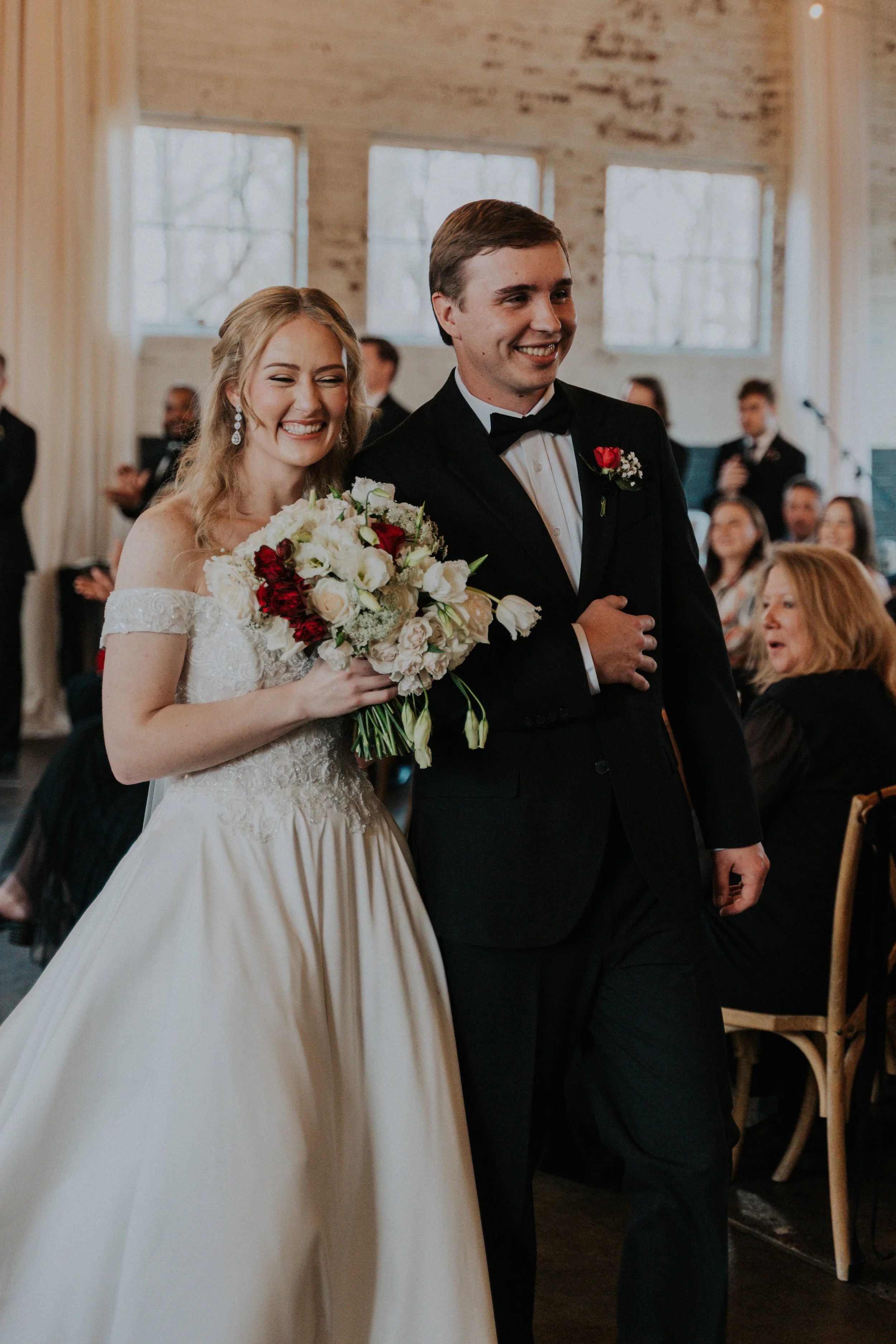 A bride and groom smiling as they walk down the aisle at a wedding ceremony, with guests in the background.