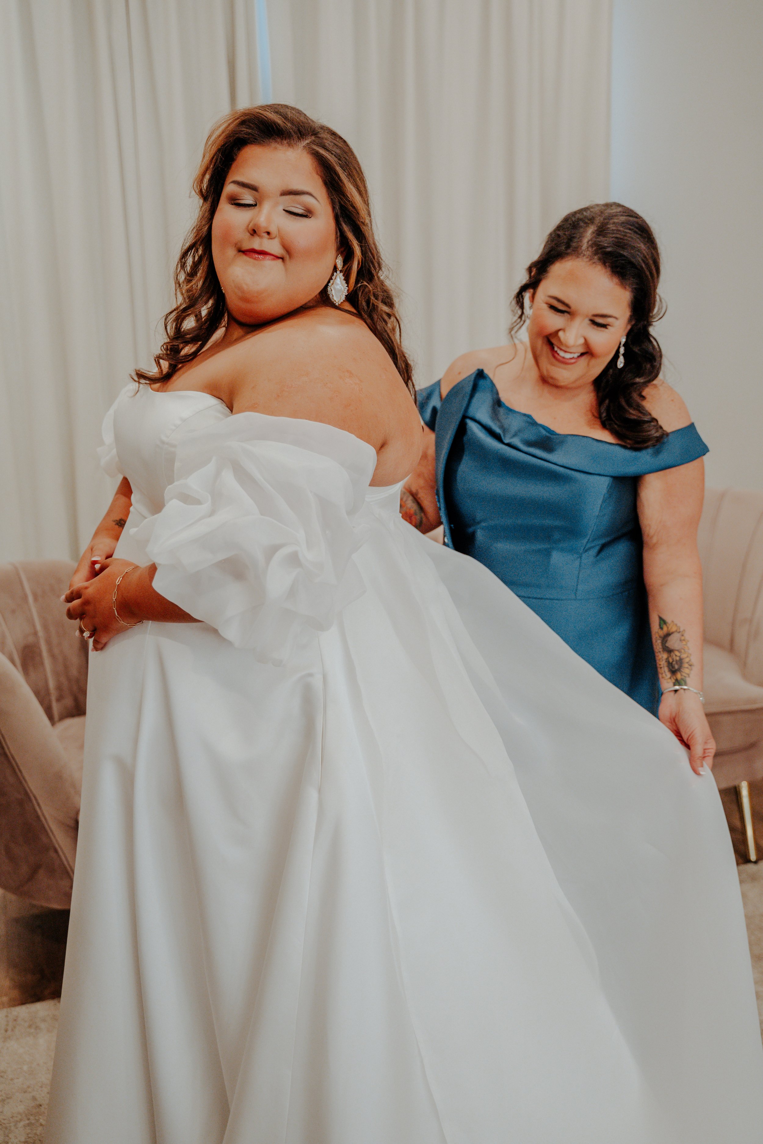 A bride in a white wedding gown with off-the-shoulder sleeves and ruffles is being assisted with her dress by a woman in a blue dress, both smiling.