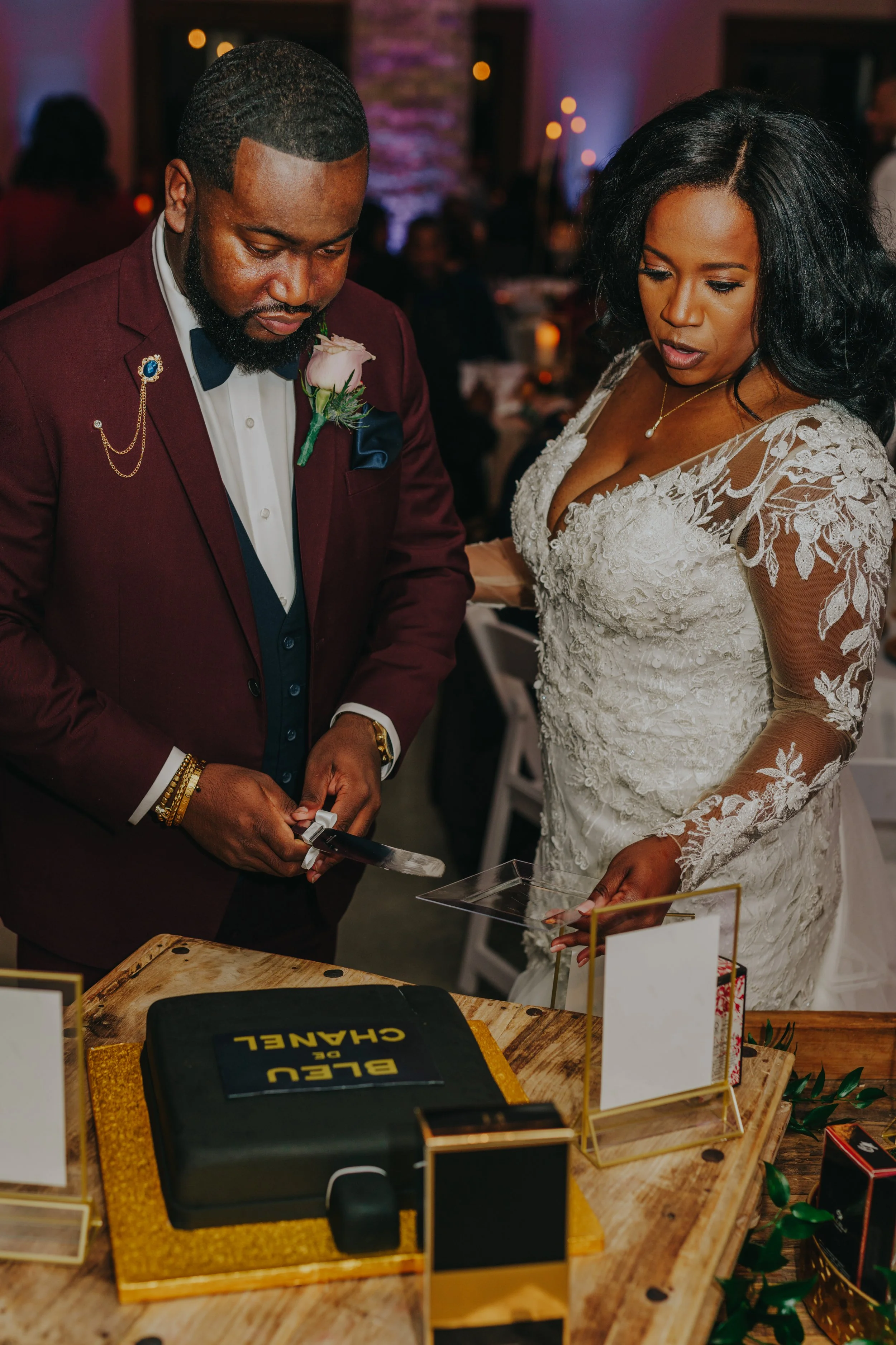 A bride and groom cutting a wedding cake at their reception, standing at a wooden table with a cake decorated in black with yellow lettering, surrounded by empty picture frames and decorations.