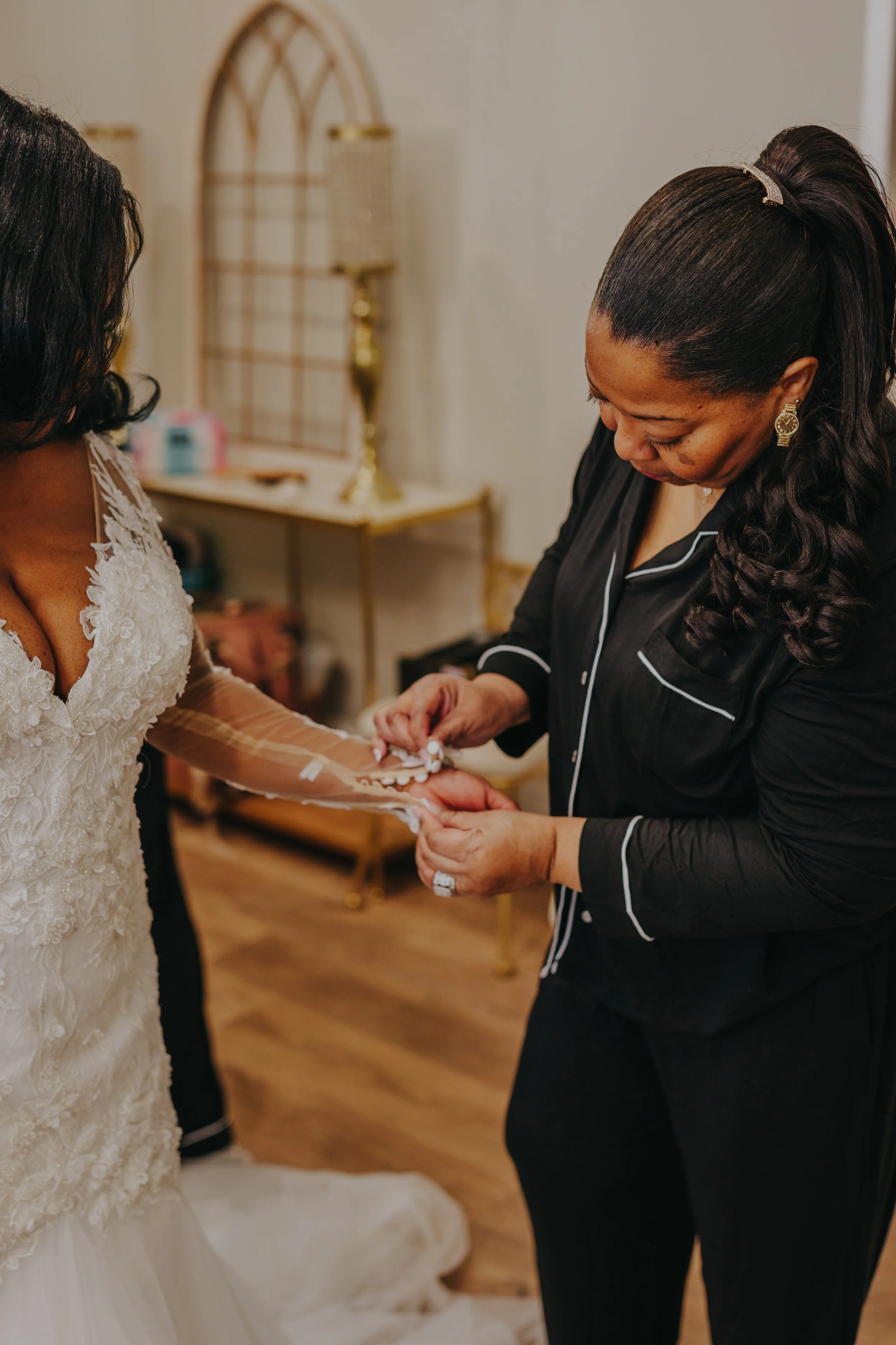 A woman in a wedding dress having her sleeve buttoned by another woman dressed in black pajamas.