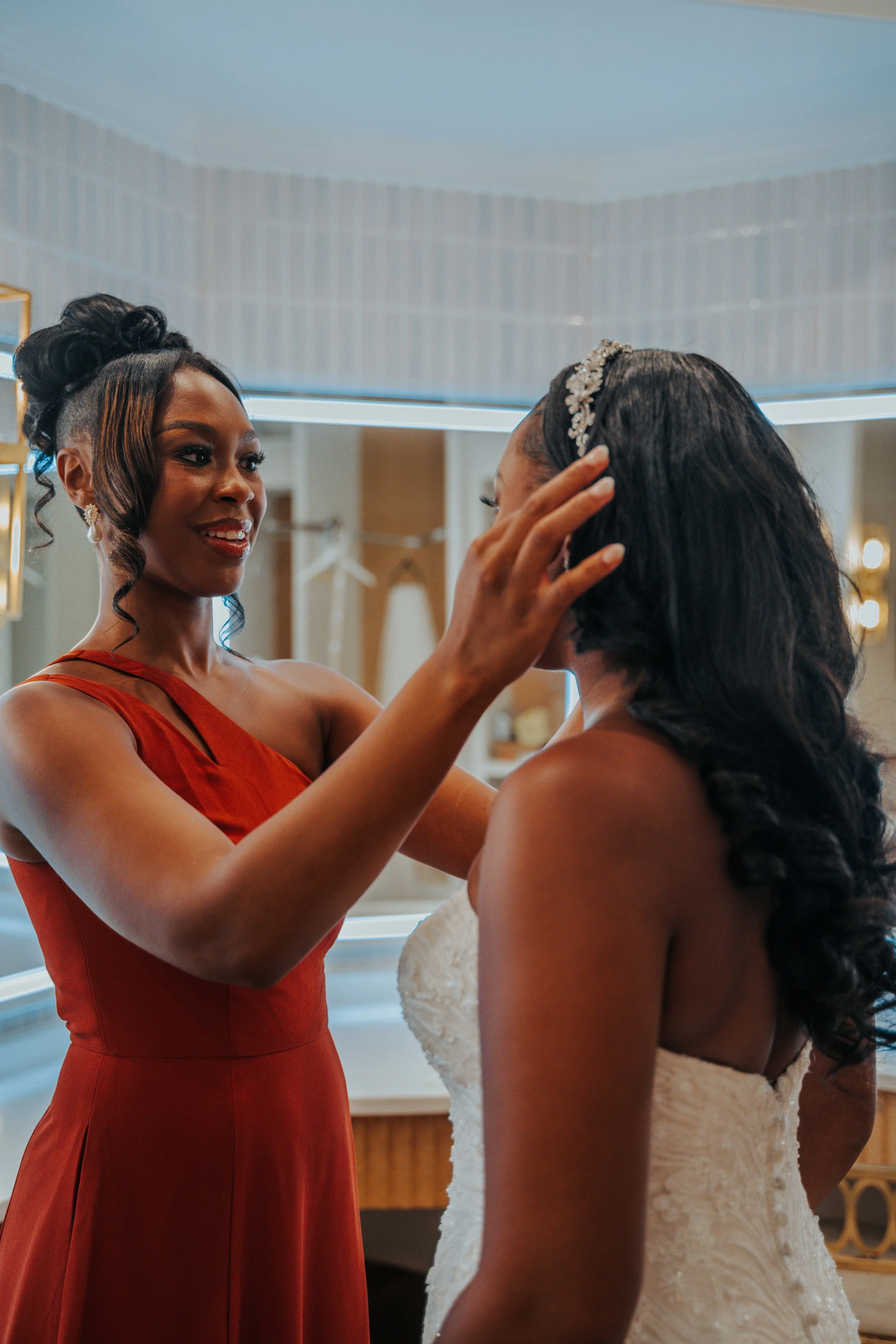 A woman in a red dress gently adjusts a bride's veil or hair in a dressing room.