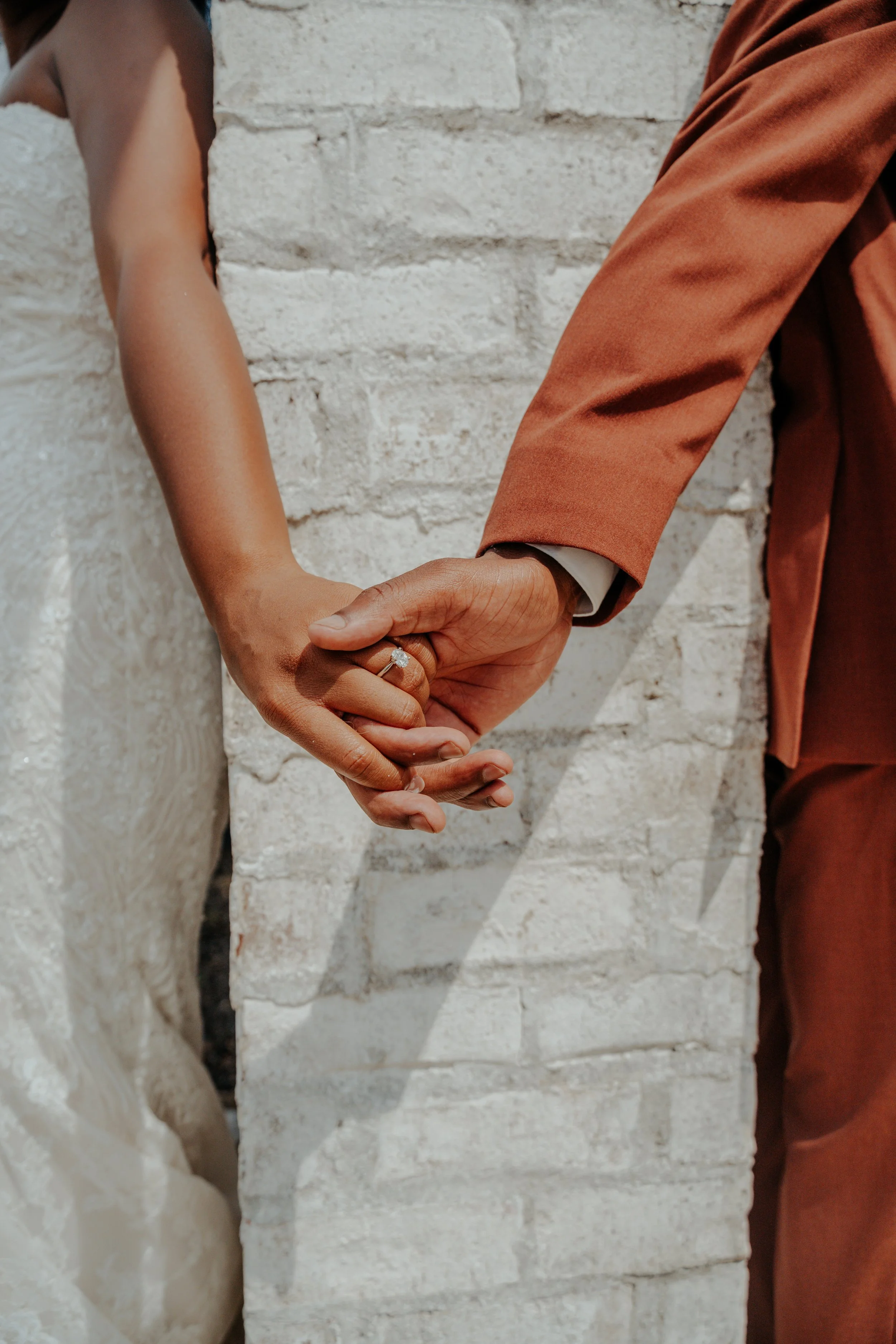 Close-up of a bride and groom holding hands, showing the wedding ring of the bride, with a white brick wall in the background.