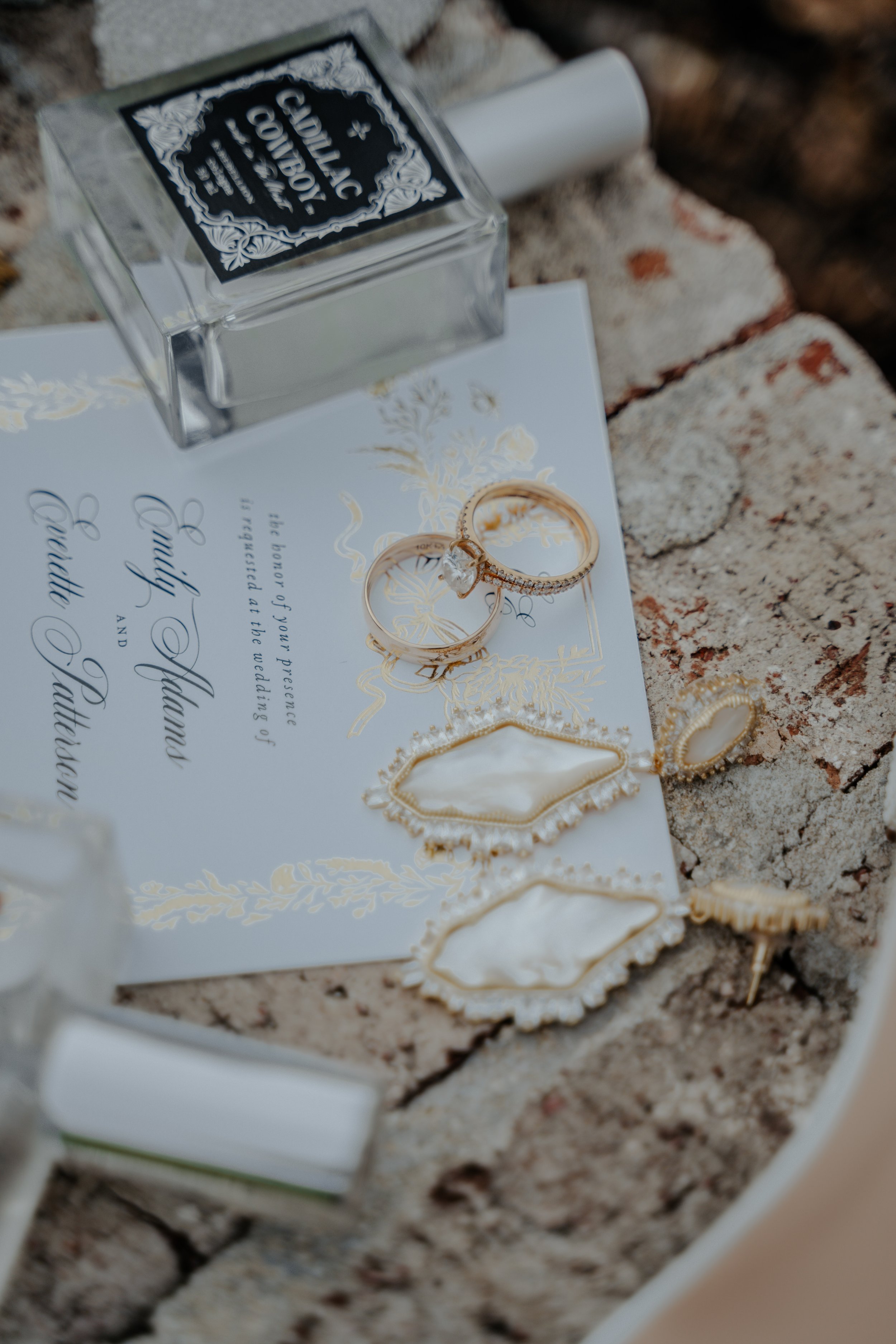 Wedding invitation with gold accents, two rings on top, and jewelry pieces around it, including earrings and brooch, on a brick surface next to a bottle of Jo Malone perfume.
