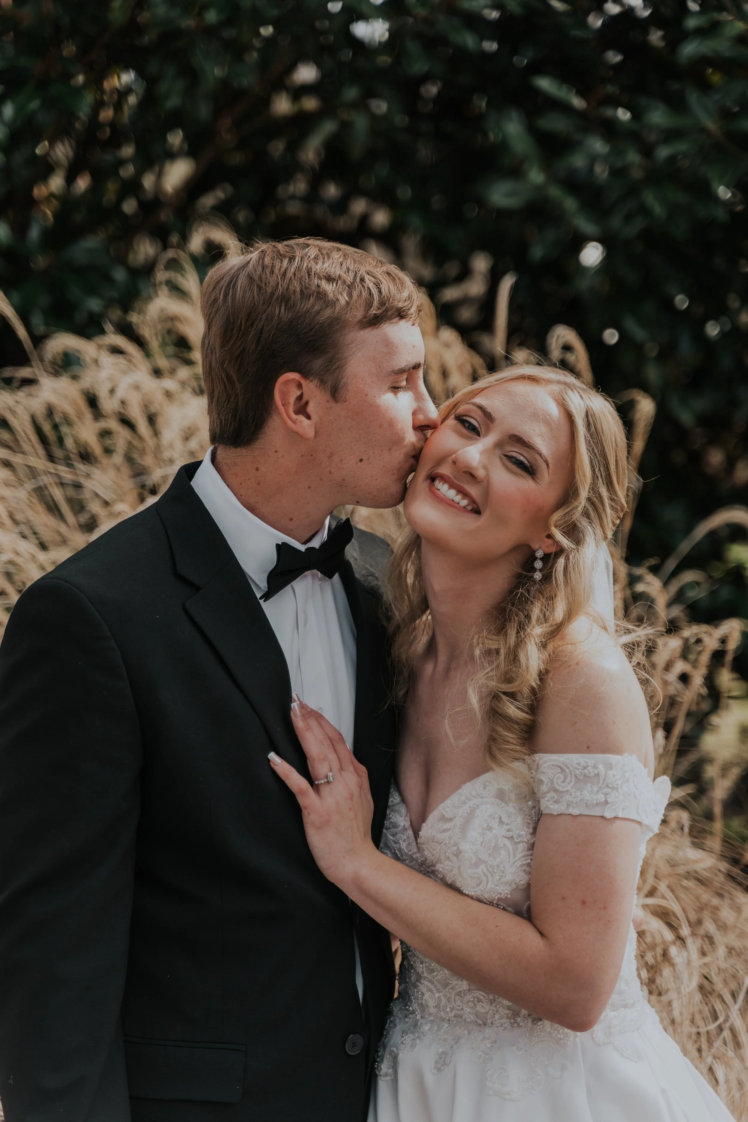 A newlywed couple, the groom in a black tuxedo and the bride in a white wedding gown, sharing a tender moment outdoors with greenery and tall grasses in the background.