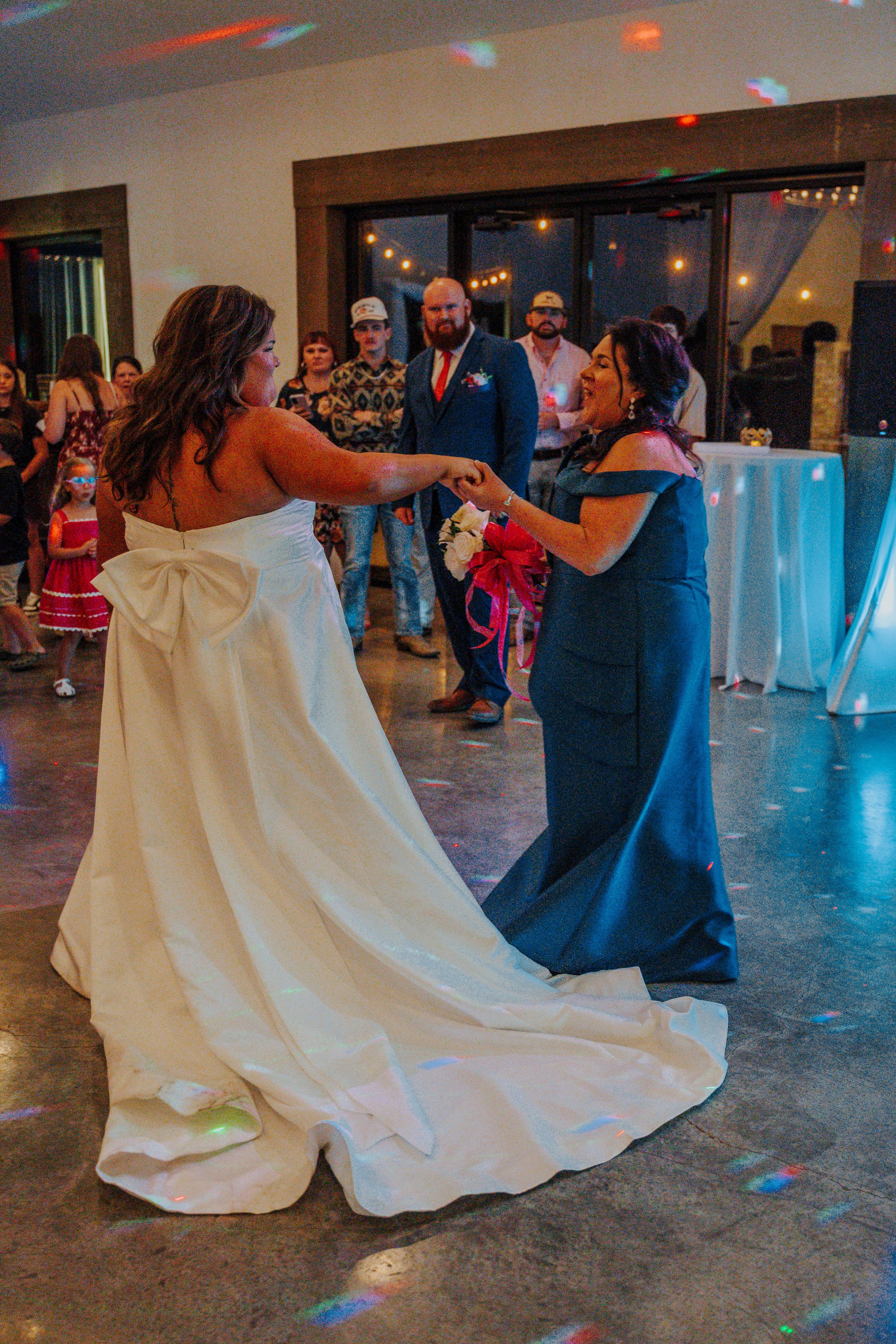 Two women dancing at a wedding reception, one in a white wedding gown with a bow, the other in a dark blue dress, with guests watching in the background.
