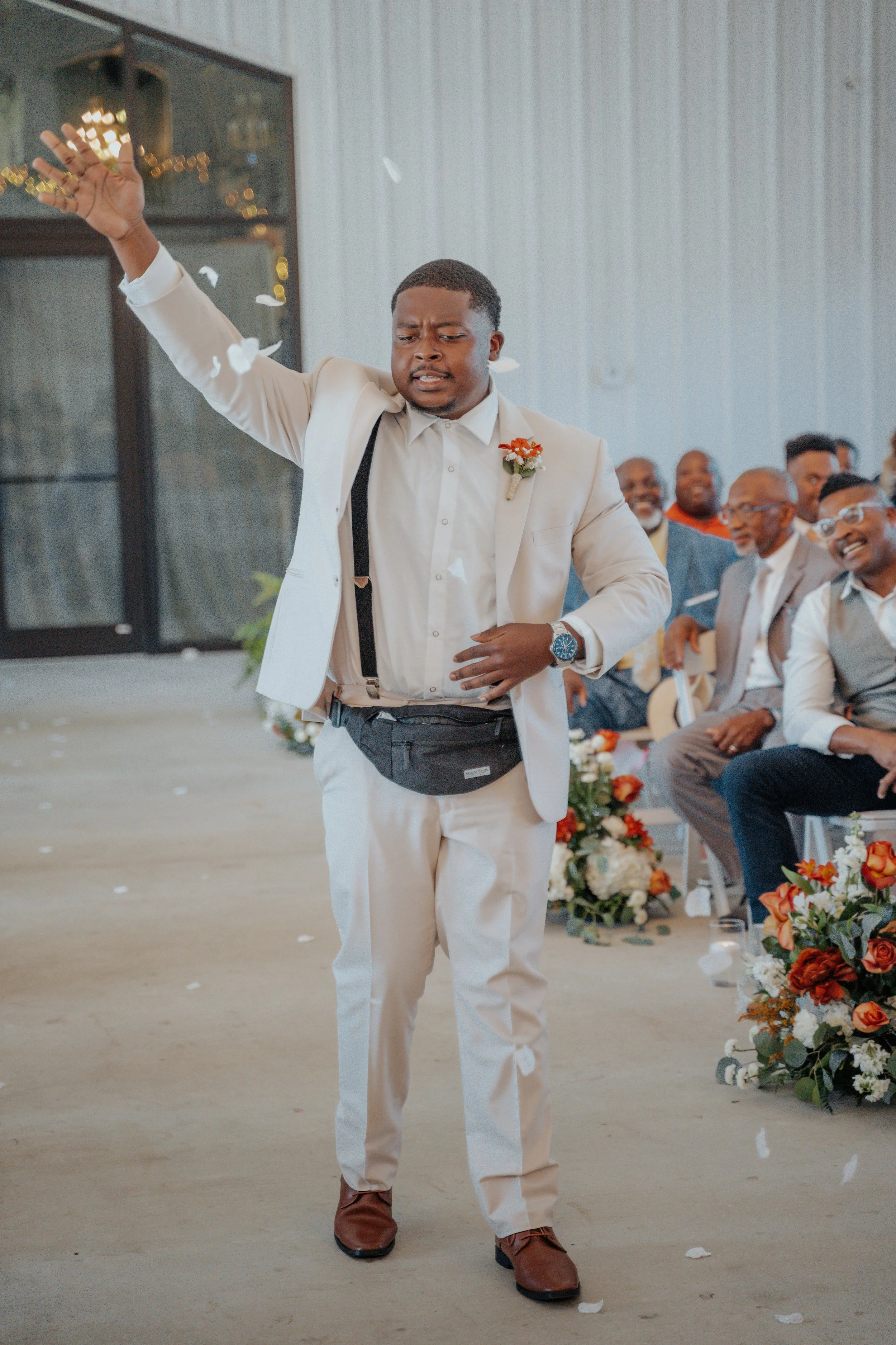 A man in a white suit and flower pin throws flower petals at a wedding ceremony. He wears a watch and a black waist pouch, with guests smiling in the background.